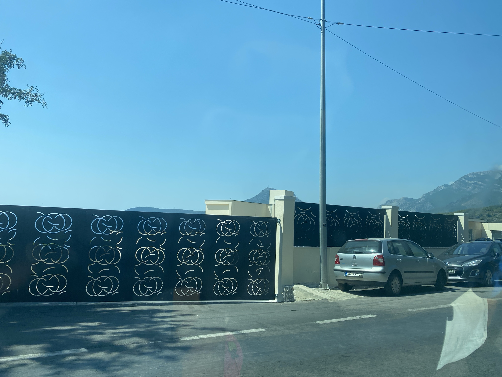 The image depicts a street scene in Sutomore, Montenegro, characterized by a large, decorative metal gate with an intricate Celtic knot pattern. The gate is black with silver cutouts and spans the width of the frame. Adjacent to the gate is a modern, cream-colored building with a flat roof and vertical pillars. In the foreground, a paved road with white lane markings is visible, with shadows cast from unseen objects. Two cars are parked on the right side of the road: a silver Volkswagen Polo and a dark-colored Peugeot. The Volkswagen Polo has a license plate with the inscription "KU AM 451". The background shows a clear blue sky, distant mountains, and a few scattered trees. The scene is captured on a sunny day.