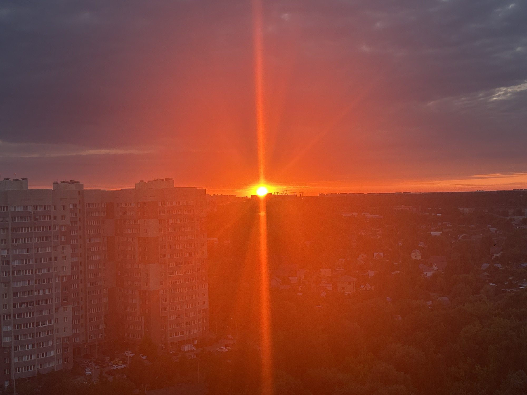 An aerial view captures a vibrant sunset over Ivanteyevka, Russia. The sky is a dramatic gradient of deep purples and fiery oranges, with the sun a blindingly bright orb near the horizon, casting a strong vertical beam of light. In the foreground to the left, the detailed facades of several tall, apartment buildings are visible, their windows reflecting the sunset. Below the buildings, a street scene with parked cars can be seen. To the right and further in the distance, a sprawling landscape of smaller houses and lush green trees creates a textured silhouette against the glowing sky. The overall impression is one of a dramatic and picturesque urban sunset.