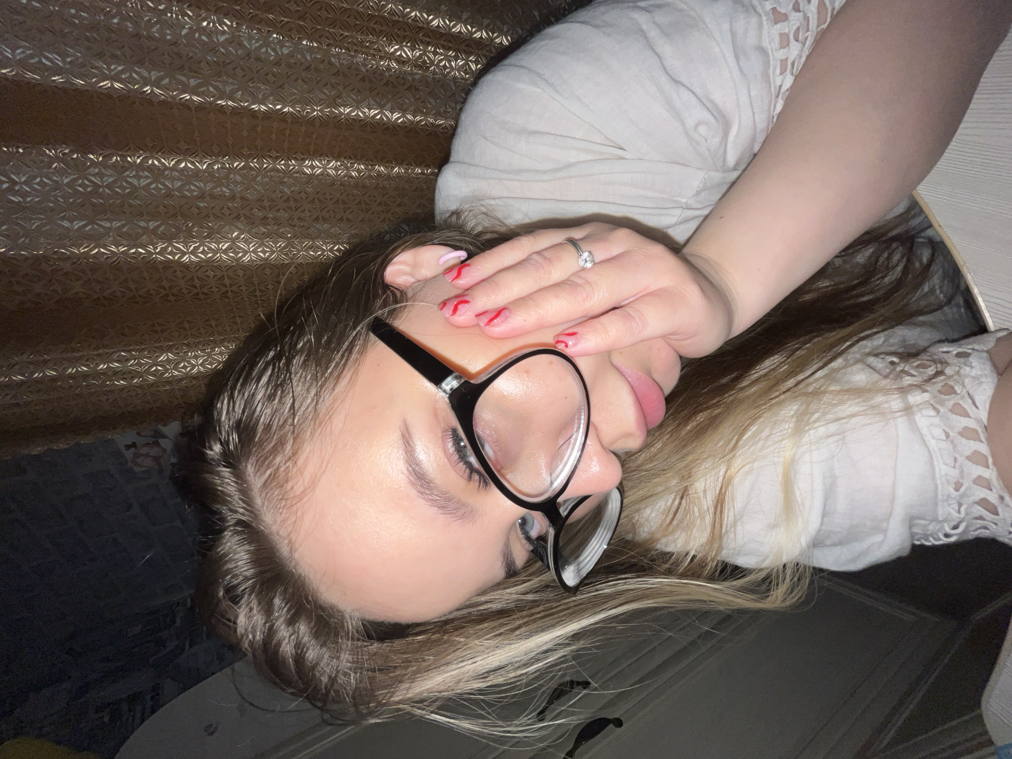 A woman lies on her side wearing glasses and a white shirt with lace detailing. Her hair is blonde and brown, and she has red nail polish with a design. She is wearing a silver ring. A textured curtain or wall covering is behind her. The location is not possible to determine from the image alone. The city of Novopokrovskaya, Russia is also not possible to determine from the image alone.
