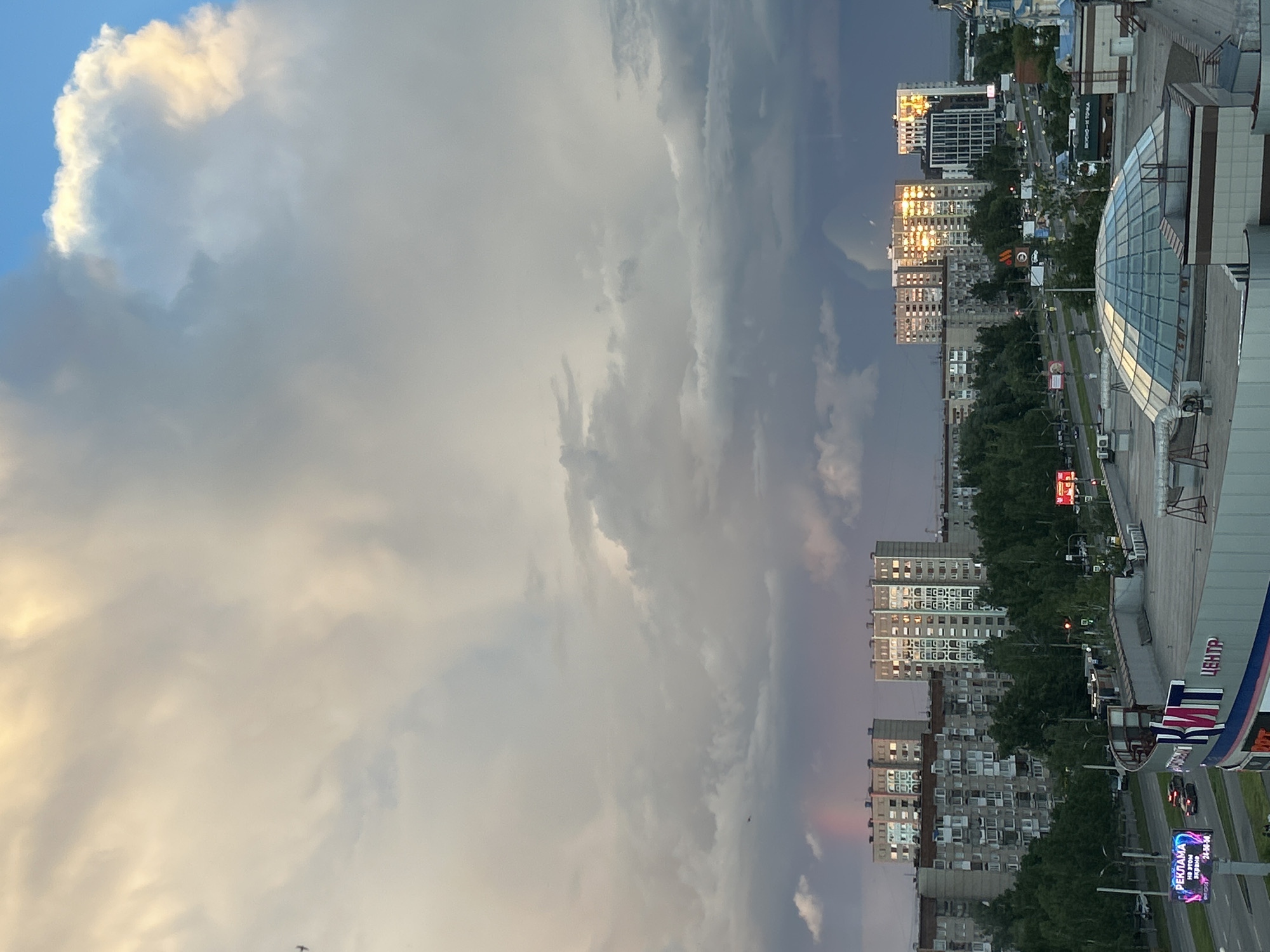 The image is an eye-level, outdoor shot showing a cityscape under a cloudy sky. The scene is dominated by billowing clouds, ranging from light grey to dark grey, indicating varying densities and potentially stormy weather. Patches of blue sky are visible between the clouds. In the foreground on the right, the edge of a building structure with a glass dome is visible. In the distance, residential buildings are seen, some reflecting sunlight. The buildings are interspersed with trees, suggesting a somewhat urban environment. Traffic is visible on the roads. A sign with Cyrillic text (ЦЕНТР) is noticeable. The location is likely Izhevsk, Russia, based on the Cyrillic script and building styles.