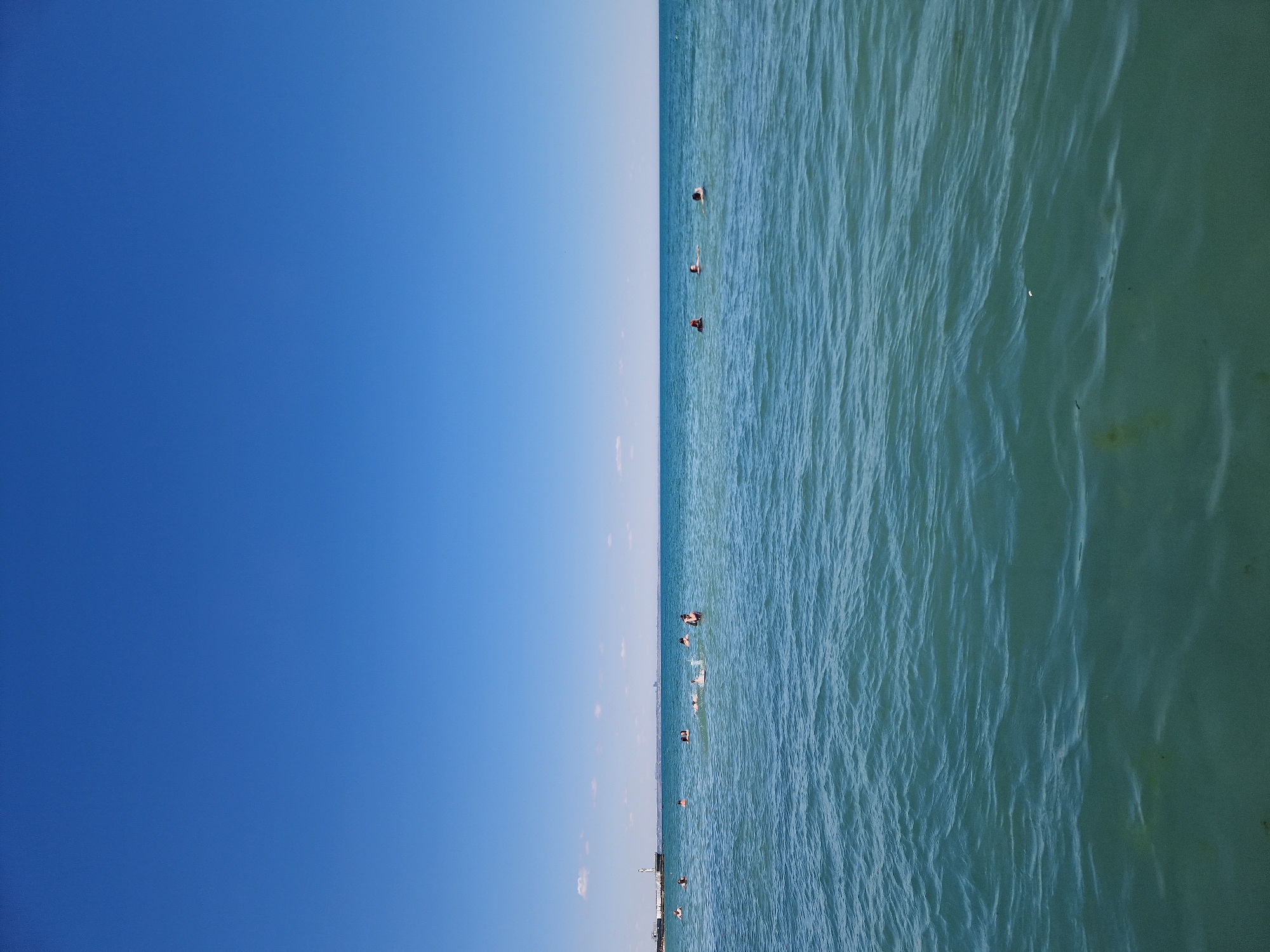 This media file displays a scene split vertically between a clear blue sky and turquoise water. The sky takes up the left half of the frame, with a subtle gradient of lighter blue towards the horizon. Scattered, wispy clouds are faintly visible in the sky.
The right half of the frame shows the water, characterized by gentle ripples and waves that create a textured surface. The water's color shifts from a brighter turquoise closer to the horizon to a darker, greener hue further into the foreground, suggesting varying depths or light conditions.
Several people are visible in the water, scattered across the right half of the image. They appear to be swimming or enjoying recreational activities in the water. Some are closer to the surface, with their heads visible, while others are more submerged. A faint, indistinct structure, possibly a pier or a distant shoreline, can be seen at the very bottom edge of the frame, along the horizon line.
The bright sky and clear water suggest a sunny day, likely during daylight hours. There are no visible signs of text in the media file. The overall mood conveyed is one of a pleasant, recreational day at a body of water under a clear sky.