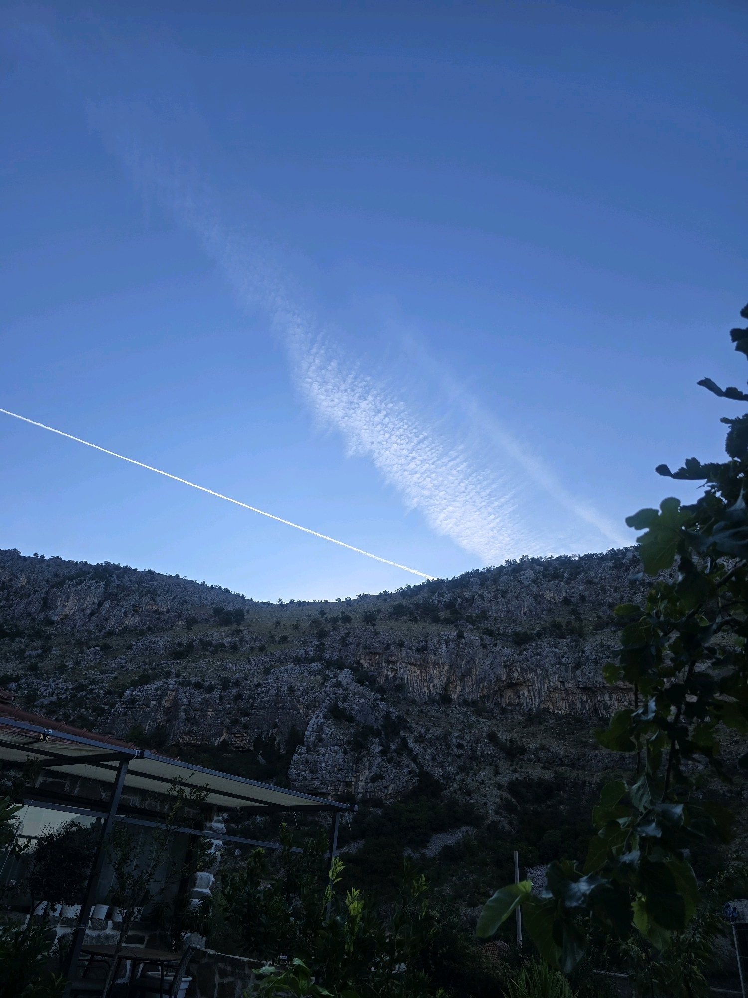 A clear, deep blue sky dominates the upper portion of the image. A single, thin white contrail stretches diagonally from the lower left towards the upper right. To the right of this contrail, a wider, more diffused cloud formation with a rippled texture is visible. The horizon is marked by a rugged, mountainous landscape. The mountains are rocky and sparsely covered with dark green vegetation, with evidence of layered rock formations and crevices.
In the foreground, to the left, a structure with a metal frame and a beige awning is partially visible. Beneath the awning, it appears to be an outdoor seating area with tables and chairs. The lower right corner of the image shows dark green foliage, possibly from a tree, with several large leaves prominent in the foreground. The overall lighting suggests either early morning or late evening. The scene is set in a natural, mountainous environment, with human elements present in the form of a covered outdoor area. The image does not contain any people or animals. The location is the city of Podgorica, Montenegro.