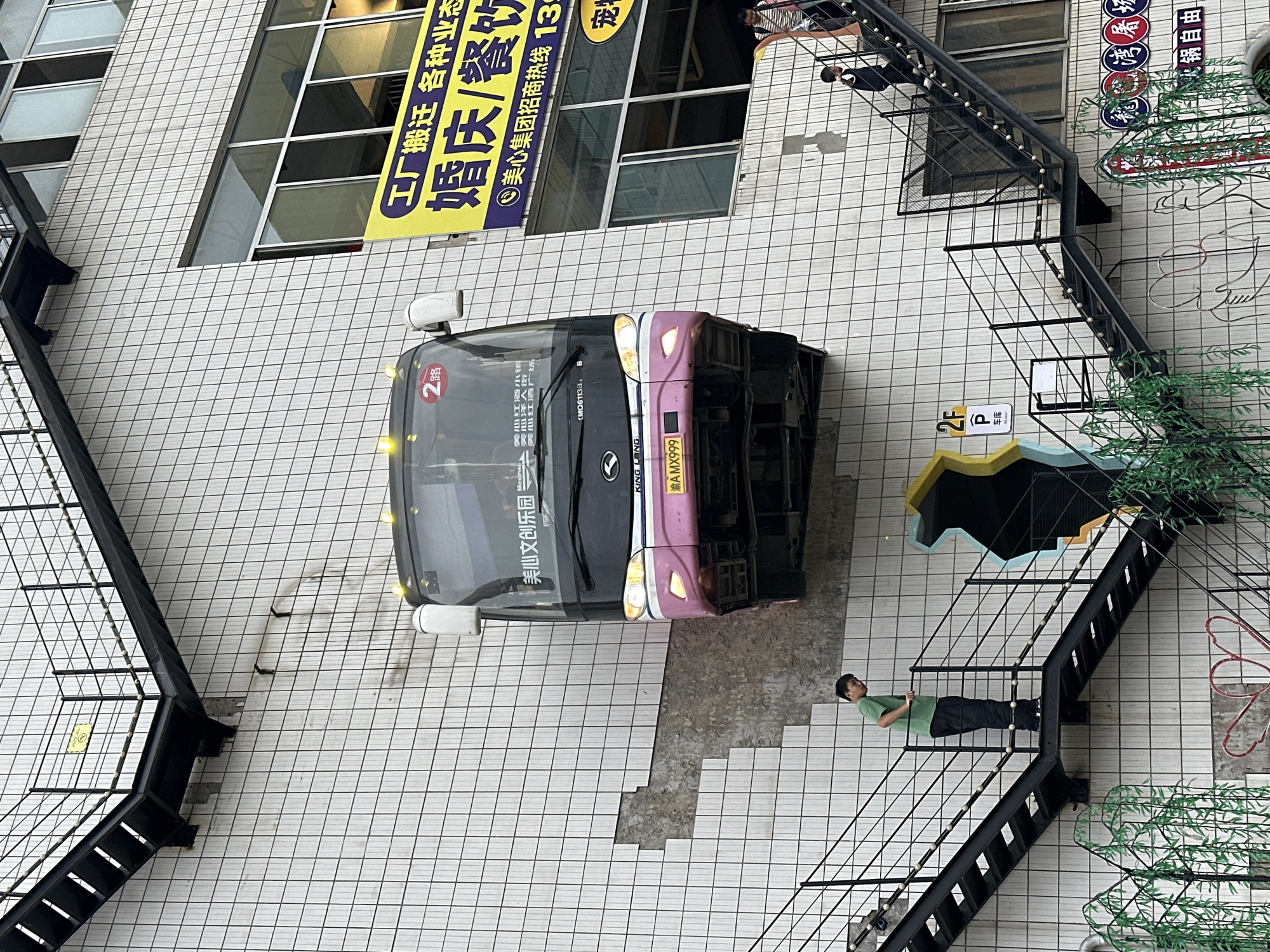 The image displays an outdoor urban scene, captured with a 90-degree counter-clockwise rotation, featuring a bus parked in front of a tiled building facade with an external staircase.
A black and pink bus is the central subject, parked on a light-colored tiled surface. The bus displays "2路" (Route 2) on its front and the license plate "渝AMX999". Text on its side windows includes "美心文创乐园" (Meixin Cultural and Creative Park), "美心红酒小镇" (Meixin Wine Town), "美心洋人街" (Meixin Foreigner Street), and "美心红酒广场" (Meixin Wine Plaza).
The background is dominated by a building with a facade covered in white square tiles. Several windows are visible, and a large yellow banner sign hangs prominently, advertising "工厂搬迁 各种业态" (Factory relocation, various business types), "婚庆/餐饮" (Wedding/Catering), "美心集团招商热线 139" (Meixin Group Investment Hotline 139), and "宠物" (Pets).
On the right side of the image, a black metal staircase with white decorative spheres on its railing ascends upwards. A man in a green t-shirt and dark pants is standing, leaning against this railing near the bottom. Further up the stairs, another person in a dark top is visible. Near the bottom of the staircase, a yellow and white sign with "2F P" and Chinese characters "车库" (parking garage) is posted. Other signage includes circular logos with "龍湾屠坛" (Longwan Butcher Shop) and "火锅自由" (Hotpot Freedom). Artificial green foliage is placed near the base of the stairs.
The scene appears to be set during the daytime, under bright, possibly overcast conditions, as evidenced by the diffused lighting and lack of harsh shadows. The location cues, including the Chinese text and specific place names like "Meixin Wine Town," suggest this is an area within Nan'an District, China, likely a commercial or entertainment complex.