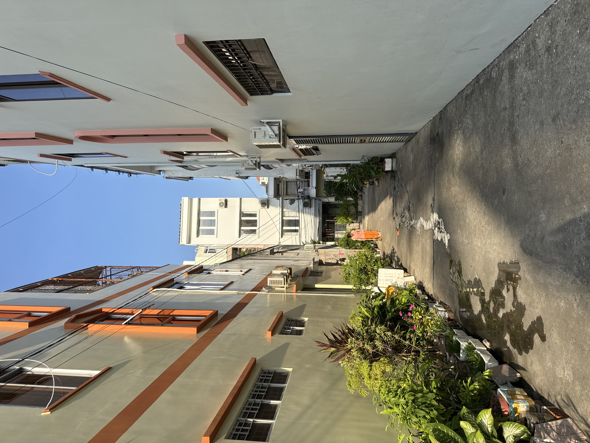 The image depicts a narrow alleyway in Nha Trang, Vietnam, flanked by buildings on either side. The building on the left is painted a pale green with orange horizontal stripes and features multiple windows. The building on the right is painted a light gray with pale pink accents around its windows. The alleyway is paved with concrete and shows signs of recent rain with several puddles reflecting the surrounding buildings and the clear blue sky.
Along the left side of the alley, an assortment of potted plants, including leafy green foliage and flowering plants, are arranged in rows. Some of the pots are simple white containers, while others are more decorative, made from patterned materials.
A person is visible in the distance, walking away from the viewer down the alley. They are wearing an orange outfit. The person appears to be alone.
In the background, several more buildings of varying heights and architectural styles are visible, suggesting a densely populated urban environment. Air conditioning units are mounted on the exterior walls of some of the buildings. Electrical wires are strung across the scene, typical of an urban setting. The overall impression is of a residential or mixed-use area within the city.