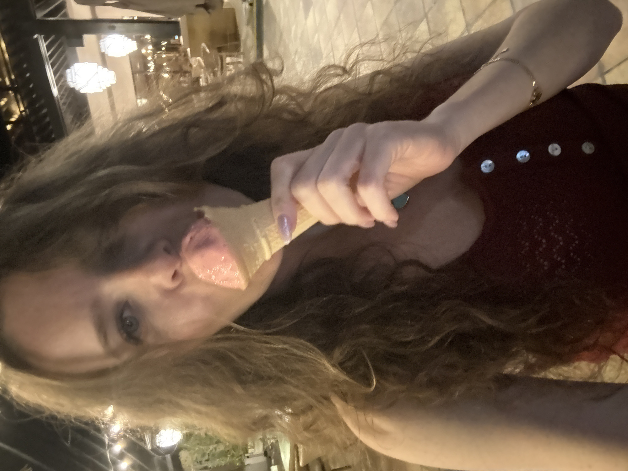 The image captures a close-up, waist-up shot of a woman eating ice cream in an outdoor, nighttime setting in Dubai, United Arab Emirates. The woman has long, wavy light brown hair that cascades over her shoulders and partially obscures the right side of her face. Her left eye, with a blue iris, is visible and directed towards the viewer. She is actively licking a scoop of pink ice cream from a light-colored waffle cone, held in her left hand. Her left fingernails are painted with a glittery pink polish. She wears a dark red, possibly maroon, sleeveless top with a textured, open-knit pattern and four visible white circular buttons aligned vertically down the front. A thin gold-colored bracelet adorns her right wrist. A small, dark blue or green circular pendant is visible hanging from a necklace on her left side, partially covered by her hair. The background is dimly lit by several artificial light sources, including white, geometric lanterns hanging from an overhead structure on the right, and blurred string lights on the far left. Structures resembling pillars and a roof, along with indistinct outdoor furniture and tiled flooring, suggest a patio or open-air dining area.