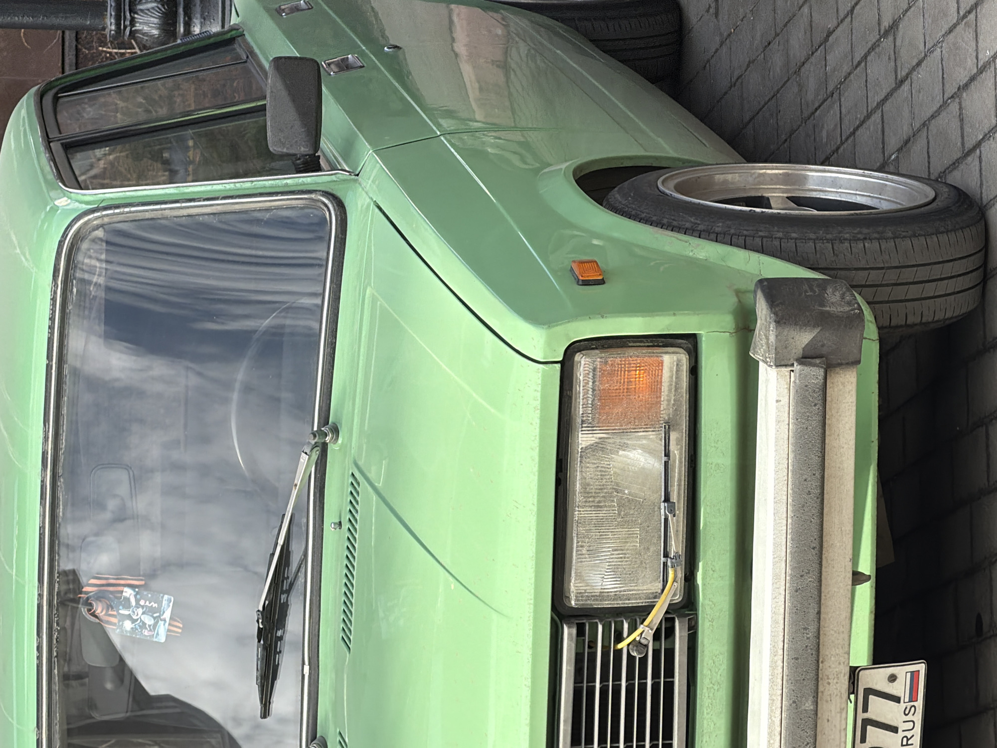 The image shows the front of a light-green vehicle, possibly a van, with a spare tire mounted on the hood. The vehicle has a boxy design with a large windshield and a chrome bumper. The license plate is visible and indicates that the vehicle is registered in Russia, specifically the 77 region, which is associated with Moscow. The background is a brick wall, suggesting the vehicle is parked near a building in an urban area, likely Moscow, Russia.