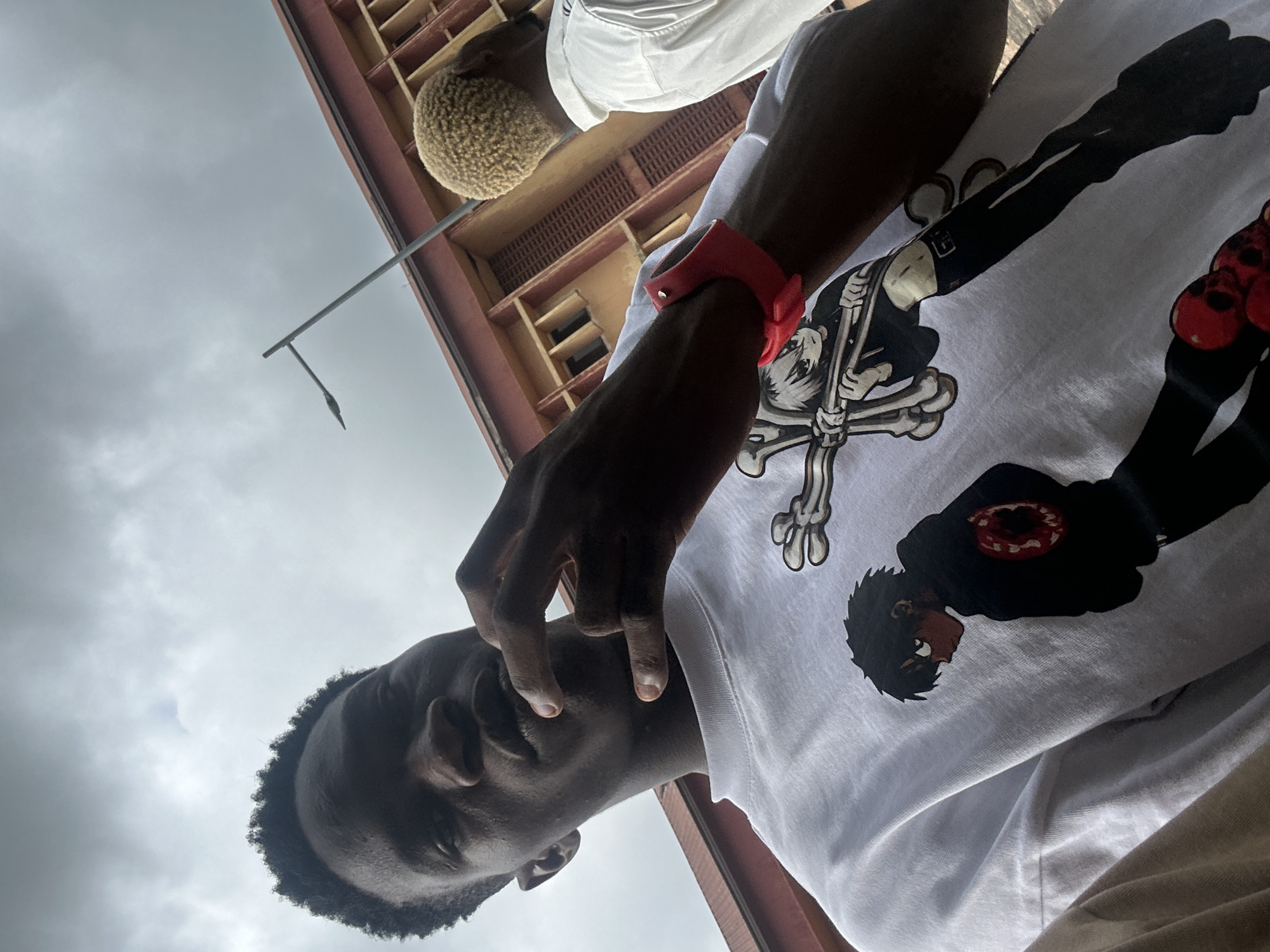 A low-angle, slightly tilted, medium shot captures a young Black man from the chest up, looking directly into the camera. He has dark, short, curly hair and his right hand is raised to his chin, with his index finger touching his lower lip. He is wearing a white t-shirt adorned with a graphic print of two anime-style characters. The character on the left is a boy with black hair and a black hoodie with a red circular design on the back. The character on the right is a girl with white hair, holding a cross made of bones, and wearing black attire. A red wristband is visible on the man's left wrist. In the background, to the right, the back of another person with short, blonde, curly hair is visible, wearing a white collared shirt. Behind them, a building with orange walls and brown trim is partially visible, featuring multiple windows. A tall, thin lamppost extends upwards into a cloudy, grey sky. The overall scene suggests an outdoor setting in a developed area, potentially a street or campus. The location of Ihiagwa, Nigeria, is provided as the context for this image.