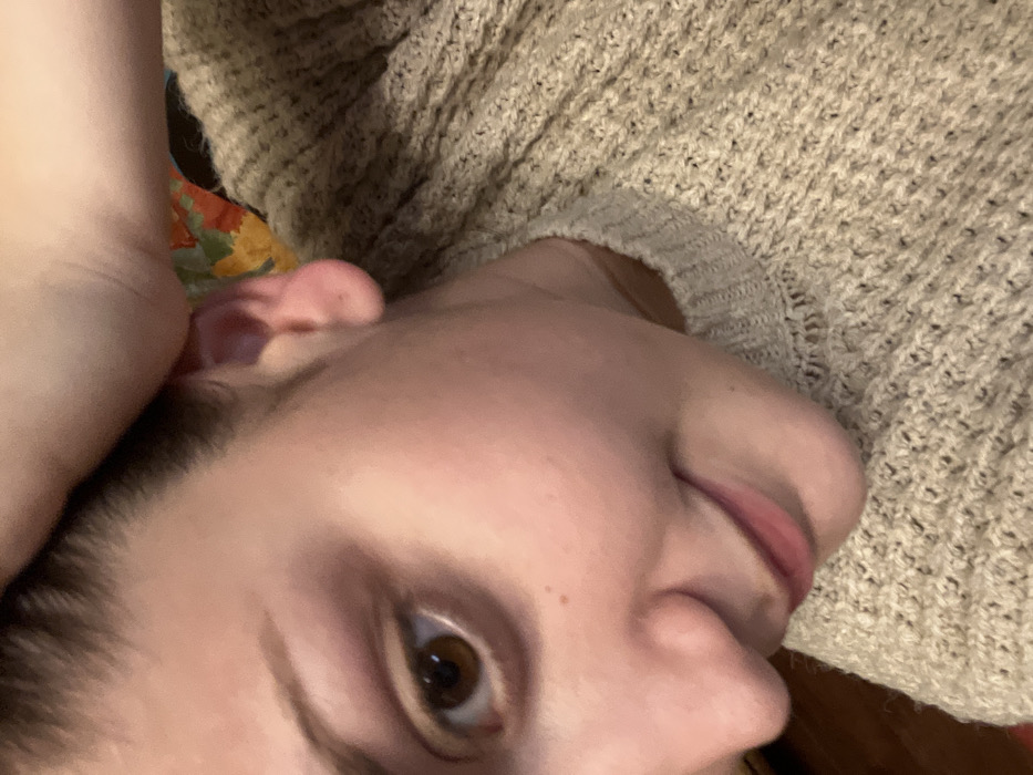 A close-up, tilted view features a person's face, with a focus on one eye, nose, and lips. The skin is fair, with visible pores and a few freckles. The person is wearing a knitted, beige sweater, which provides a soft, textured background to the upper right. Another arm is visible above the ear on the left side of the image. The overall composition is intimate, highlighting the details of the person's complexion.