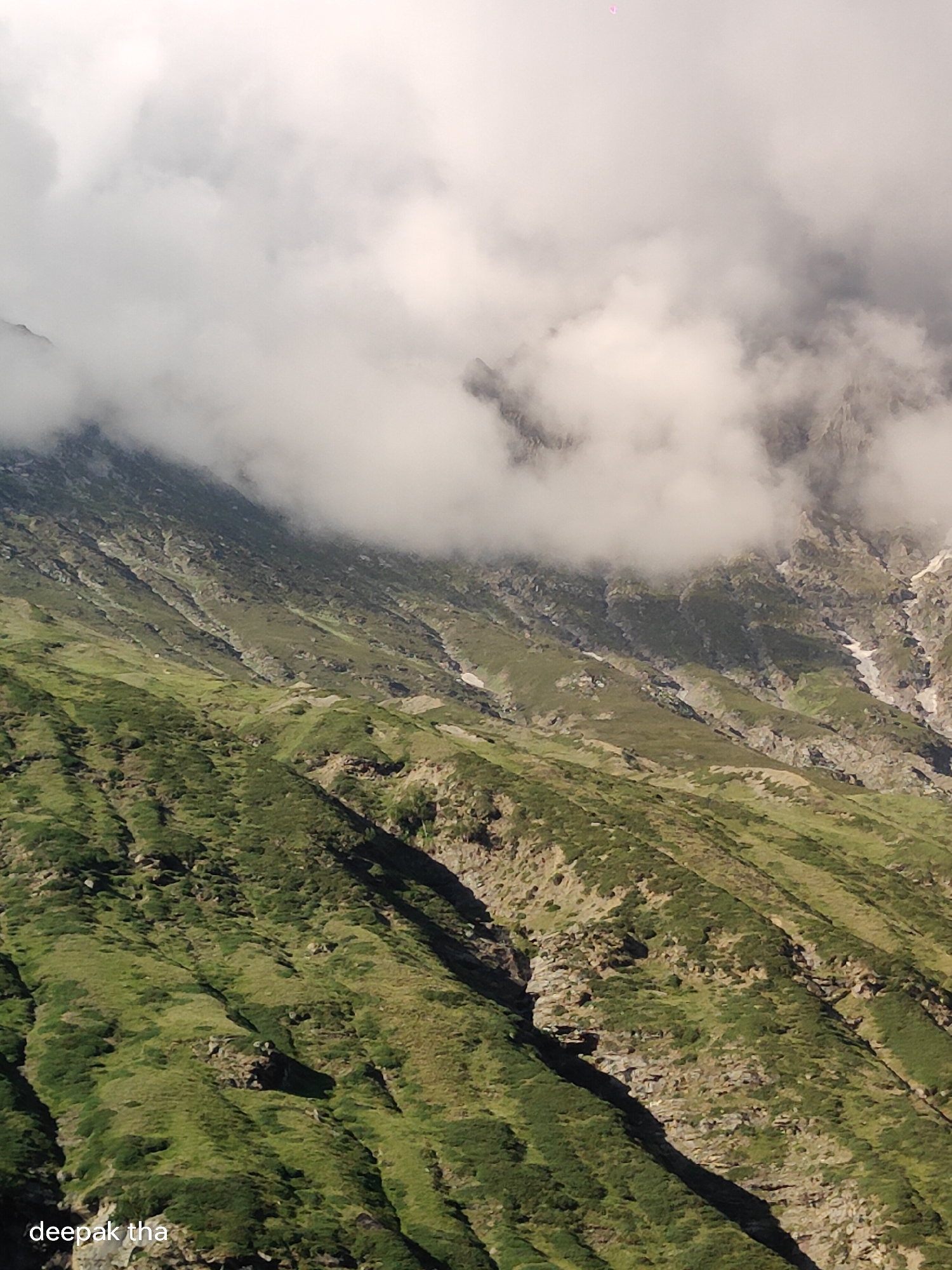 The image shows a mountainous landscape in Khangsar, India. The terrain is steep and covered in green vegetation, with rocky outcrops and patches of snow visible. Heavy clouds partially obscure the upper reaches of the mountains, creating a dramatic and atmospheric scene. The lower left corner of the image contains a watermark with the text "deepak tha".