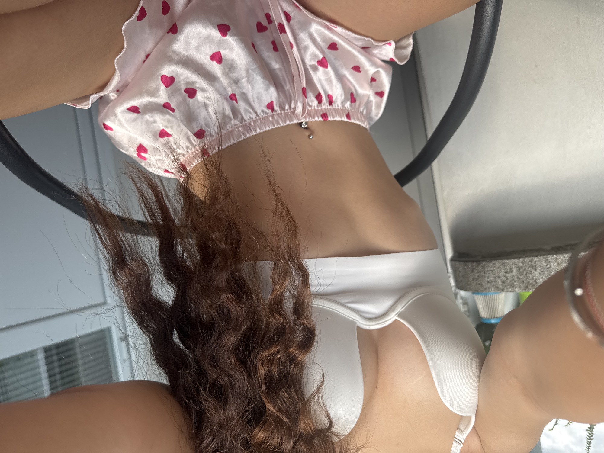 The image shows a close-up of a person from the chest down, sitting on a chair outdoors. The person is wearing a white bra and pink silky shorts with a heart pattern. Their belly button is pierced with a silver belly ring. Their hair is long and curly brown. The background appears to be a balcony or patio with a railing, a wall, and some outdoor furniture. No specific location in Plovdiv, Bulgaria, can be determined from the image.