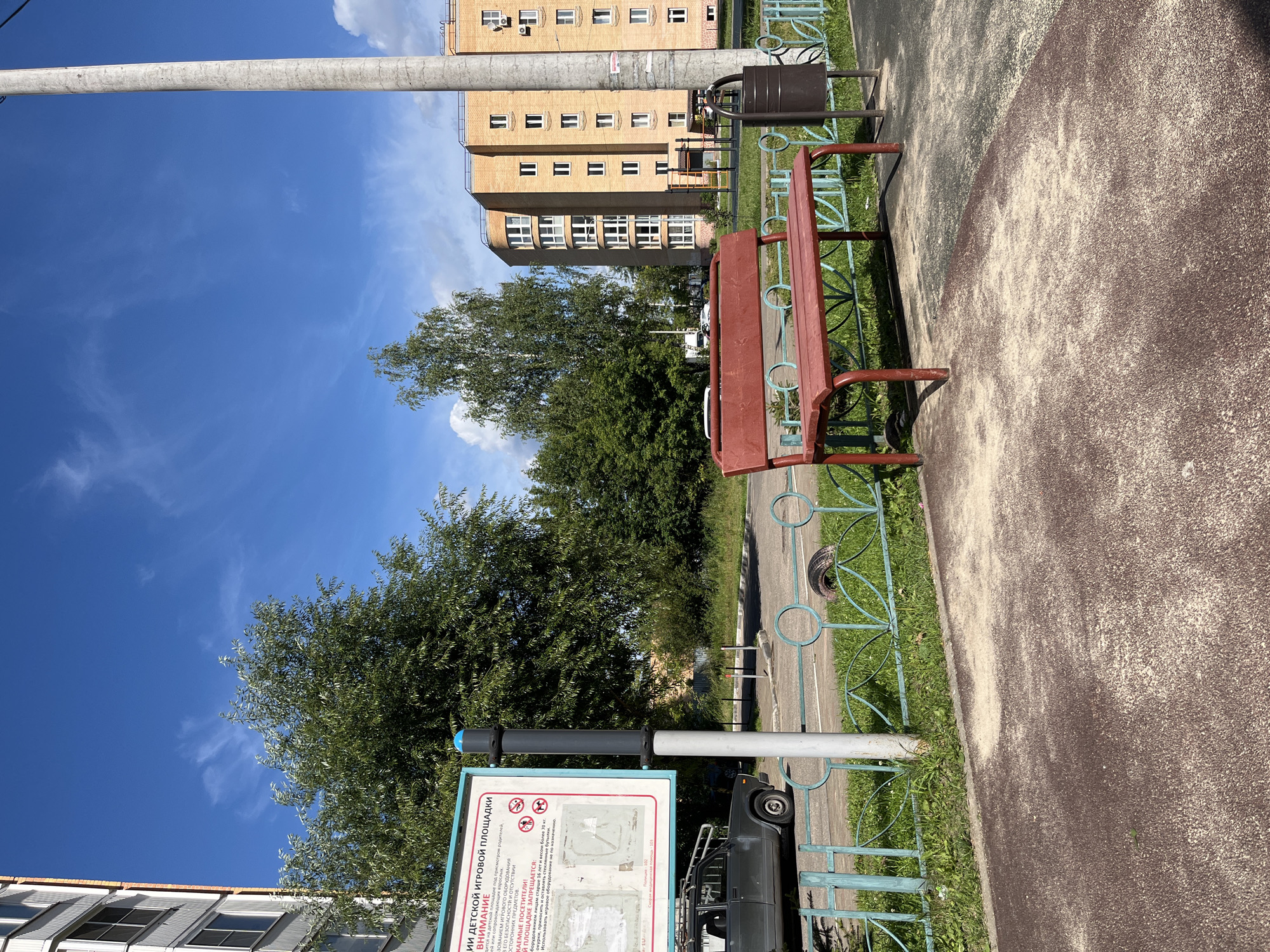 The image depicts a public area in Balashikha, Russia, on a bright, sunny day with a clear blue sky. In the foreground, a red metal park bench with slatted seating and backrest is positioned on a sandy surface. To the right of the bench, a dark-colored trash receptacle stands next to a light-colored concrete pole. A turquoise metal fence with a decorative pattern runs horizontally across the mid-ground, separating the paved area from a grassy strip. Behind the fence, a dark-colored car is partially visible, parked on a road.
In the background, a multi-story residential building with a yellow brick facade and numerous windows dominates the right side of the frame. Adjacent to it, a cluster of green trees creates a natural backdrop. A tall, slender concrete pole is prominent on the right side of the image, extending vertically from bottom to top. A sign, partially obscured by the concrete pole, is mounted on a metal post to the left of the bench. This sign features text and illustrations, likely providing information or rules for the area. The overall scene suggests a communal outdoor space, possibly a playground or park area within a residential district.