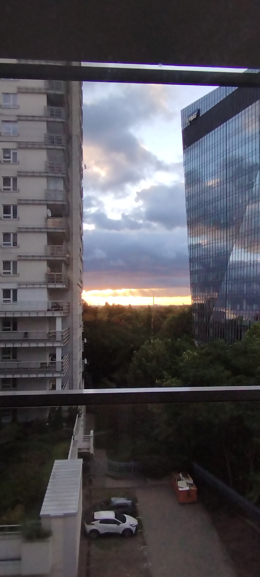 This image captures a view from a balcony overlooking a city scene at dusk, likely in Warsaw, Poland. The prominent features include a tall, light-colored residential building on the left, characterized by multiple balconies and windows. To the right, a modern, glass-facaded skyscraper stands, reflecting the dramatic cloudy sky. The sky itself displays the warm hues of sunset breaking through dark, dramatic clouds. Below the buildings, a dense area of trees forms a green canopy. In the foreground, a dark metal railing is visible, suggesting the vantage point. Further down, a parking area is visible with two vehicles, one white SUV and another darker car, and an orange dumpster. The overall impression is an urban landscape transitioning from day to night, with a mix of architectural styles and natural elements.