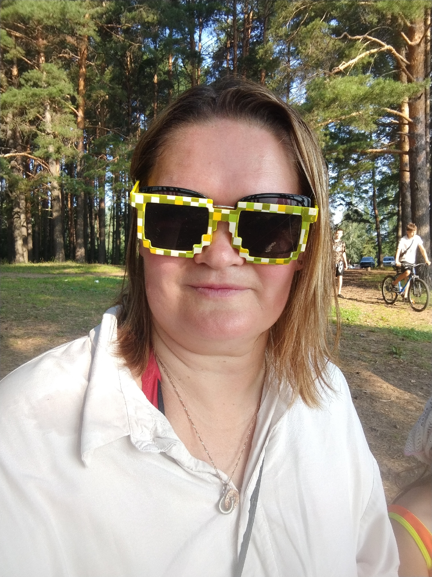 In this medium shot, a woman with shoulder-length blonde hair is the central subject, wearing distinctive pixelated sunglasses with a yellow and green checkered pattern and a white collared shirt. She is positioned in the foreground, facing the camera with a neutral expression. A thin, delicate necklace with a circular pendant is visible around her neck.
Behind her, the scene unfolds in a wooded area with tall pine trees and a grassy clearing. The sunlight filters through the trees, suggesting it is daytime, likely late afternoon or early evening given the warm lighting. In the background, two young people are visible. One, a person in a patterned t-shirt and shorts, stands looking towards the right of the frame. Further to the right, another individual rides a bicycle. Several parked cars are also discernible in the middle ground, hinting at a public park or recreational area.
The overall atmosphere appears relaxed and casual, possibly a day out in nature. The setting suggests an outdoor recreational space, common in areas like Chaikovsky, Russia, during warmer months. No text is visible in the image.