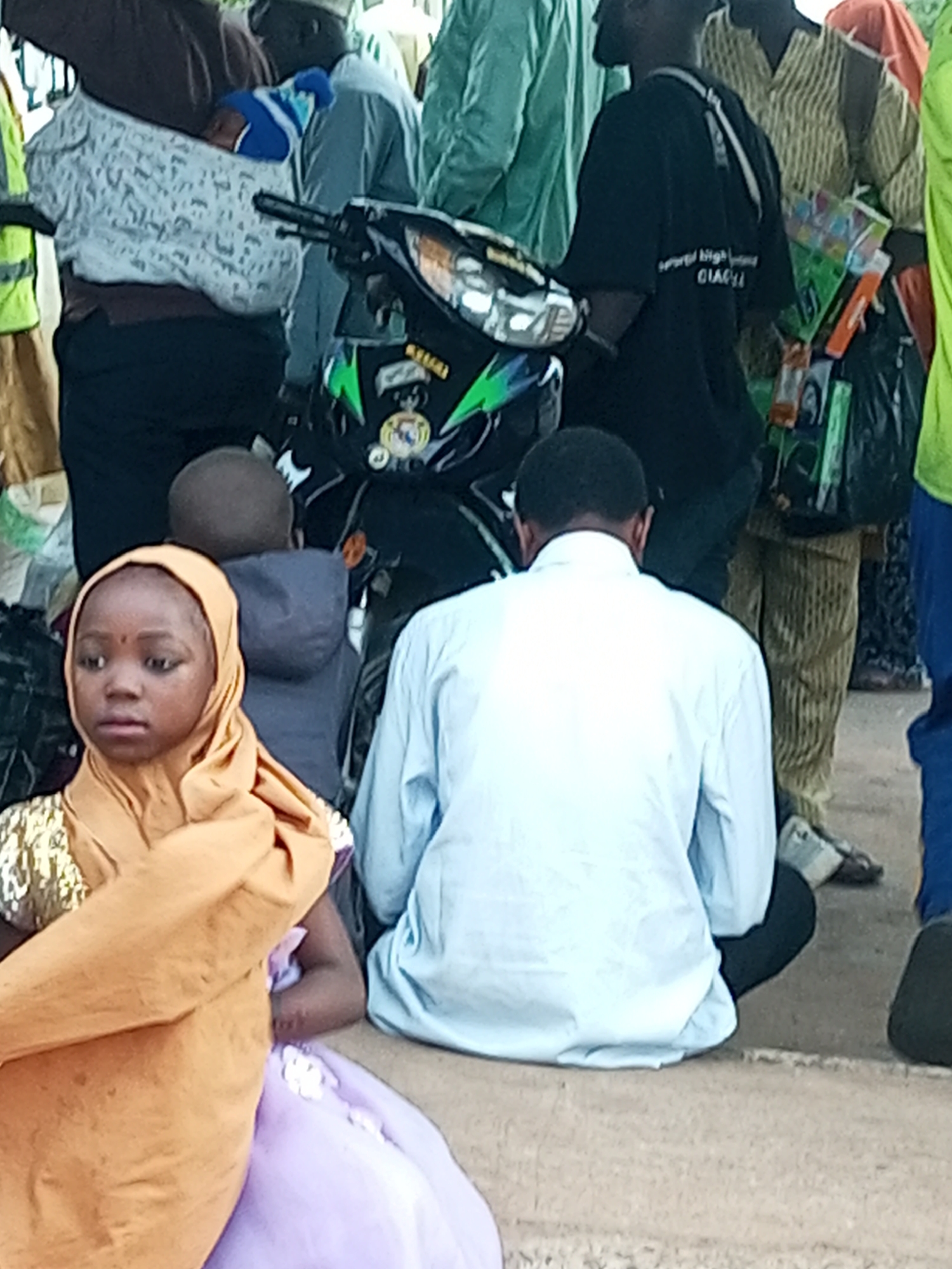 A group of people, including children and adults, are gathered around a motorcycle in the city of Gombe, Nigeria. A young girl in an orange hijab and a purple dress is in the foreground, looking to her left. Behind her, a person wearing a light blue shirt sits facing away from the camera. Other individuals, some in traditional attire, are visible in the background, along with a motorcycle adorned with colorful decals. The ground appears to be a dusty or sandy surface.