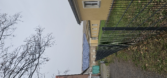 The image displays an outdoor scene in Kamenz - Kamjenc, Germany, under an overcast sky, indicating late autumn or winter. In the foreground, a dark grey gravel path runs along the left, adjacent to a black metal grid fence extending from the lower right towards the mid-ground. The ground by the fence shows sparse grass and scattered brown fallen leaves. A dark-colored bicycle is parked, leaning against the fence on the gravel path side, near a teal-colored metal container. To the far left, a portion of a red-brick building is visible. Behind the fence, on the right, a long, single-story building with a light yellow facade and several white-framed rectangular windows is present. In the background, a large building features a roof entirely covered with numerous blue solar panels. Additional long, yellow-colored buildings with similar architecture are visible behind the initial yellow structure. No individuals are present in the scene.