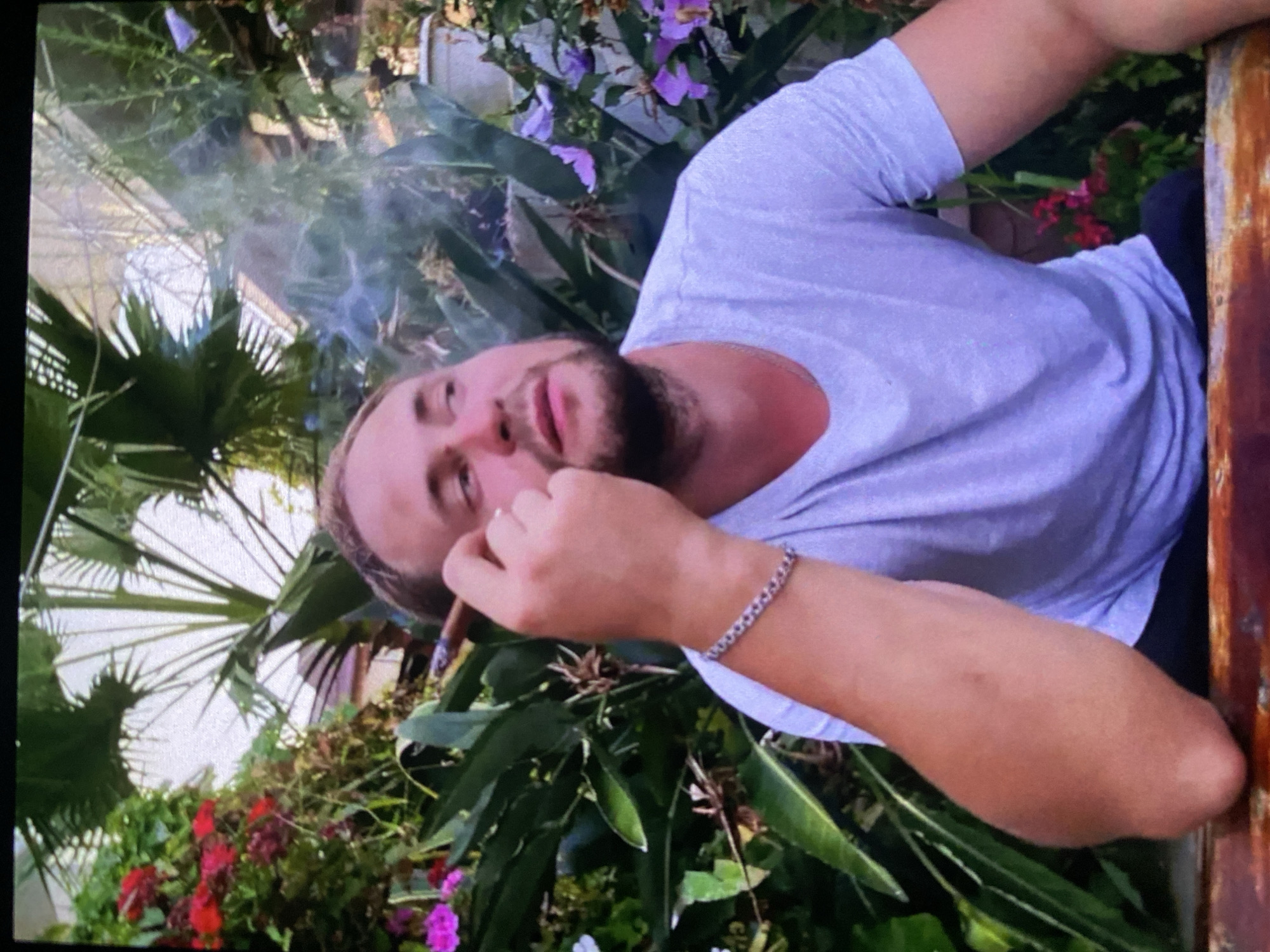 A man with short hair and a beard is smoking a cigar outdoors, surrounded by lush greenery and flowers. He is wearing a light-colored t-shirt and a bracelet. The location appears to be a greenhouse or botanical garden. There is no indication of the location being Kazan, Russia.