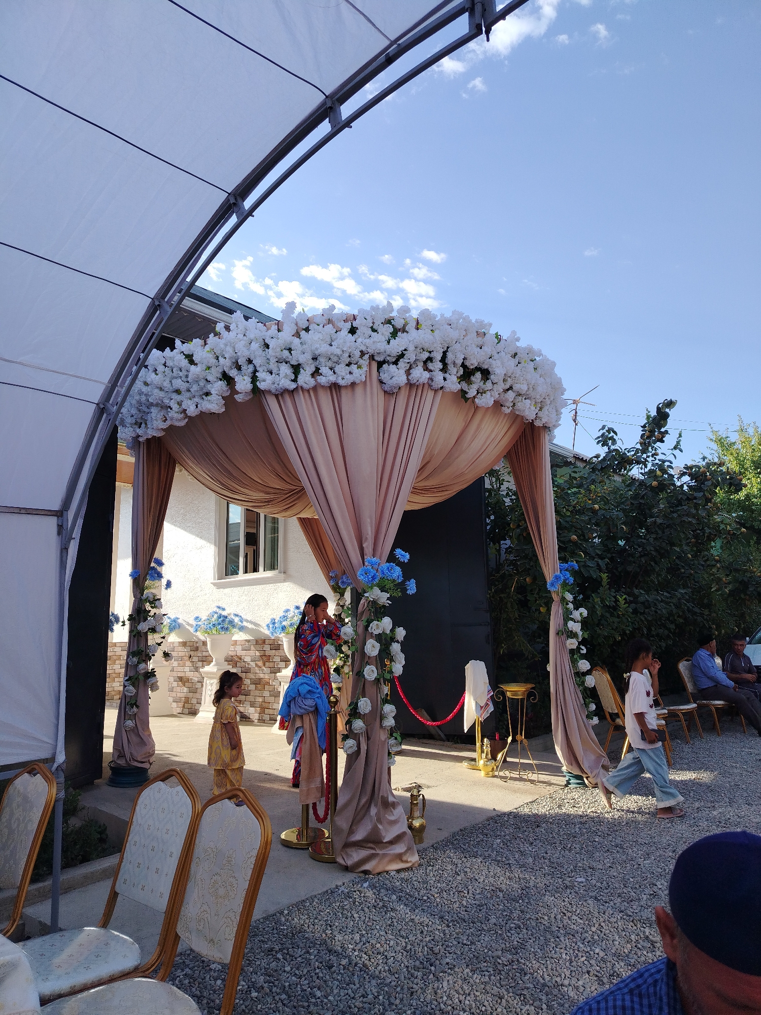 This image depicts an outdoor event setup, likely a wedding or celebration, under a large white tent. The centerpiece is a decorated archway with draped fabric in a champagne color, adorned with numerous white flowers at the top and smaller blue and white floral arrangements cascading down the sides. Several chairs with ornate gold frames and patterned cream upholstery are arranged in rows, some in the foreground and others further back.
In the middle ground, a woman in a brightly colored patterned dress stands near the archway. A young girl in a yellow outfit is positioned to her left, and another child in a white shirt and blue jeans walks across the gravel ground in the right midground. In the background, a white building with windows is visible, along with trees and other foliage. Several adults are seated on chairs in the far right background, some facing away from the camera. The ground is covered in small, light-colored gravel. The sky above is clear blue with scattered white clouds. The overall impression is that of a festive outdoor gathering.