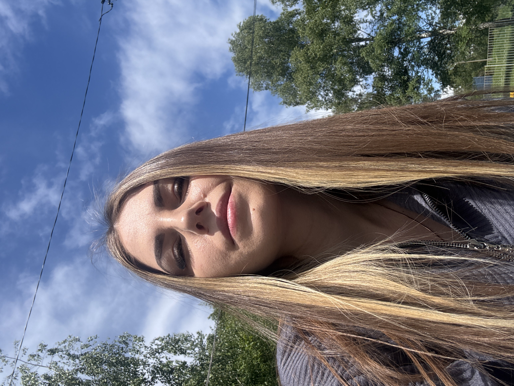 A young woman with long blonde hair is pictured in a close-up selfie outdoors on a sunny day. Her face is angled towards the camera with a slight smile. She is wearing a grey zippered garment. Behind her, there is a blue sky with scattered clouds and green trees, possibly in a park or wooded area. A utility pole or wire is also visible extending into the sky. The location, Shevlyakovo, Russia, cannot be verified from the image.