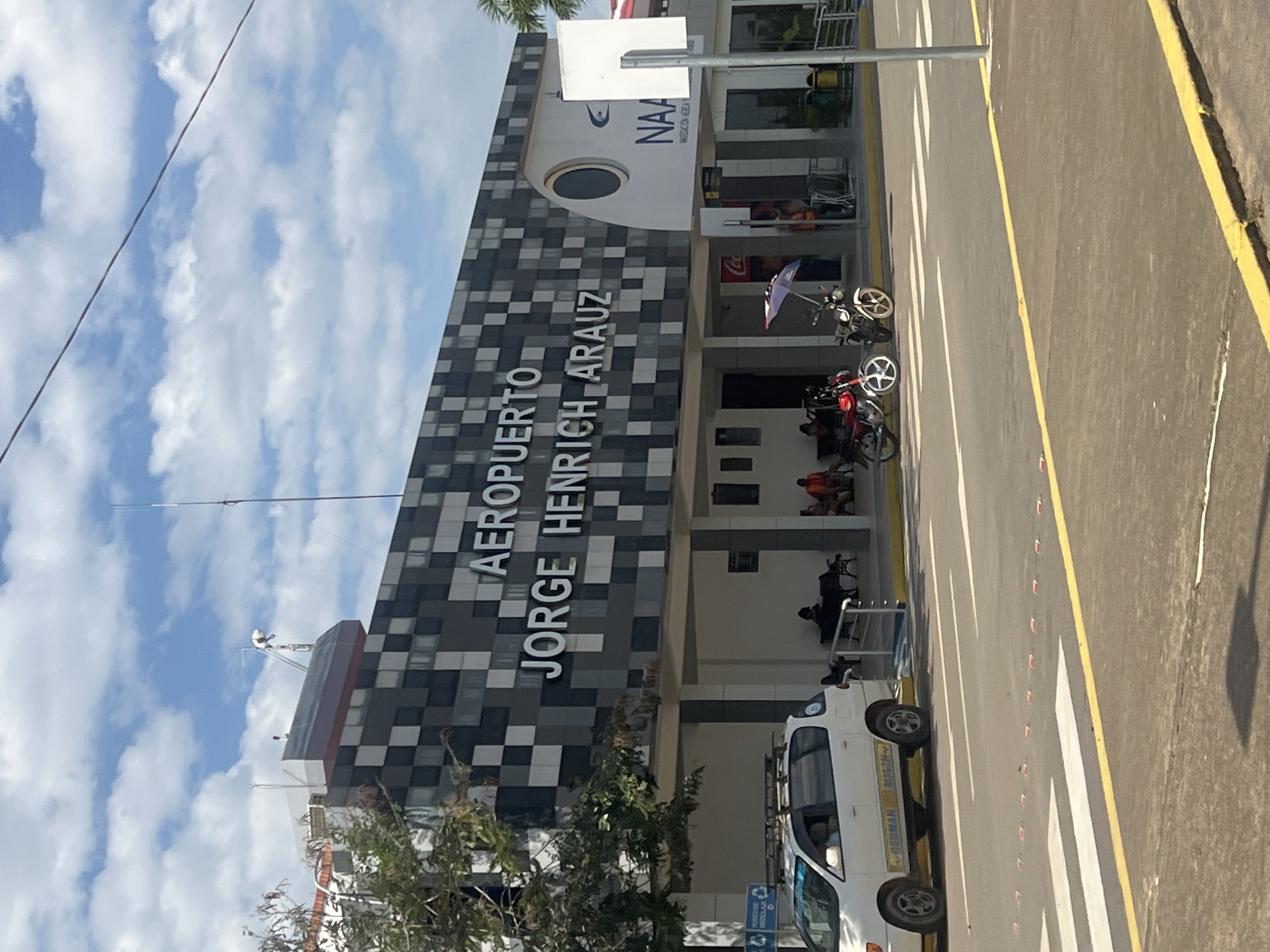 The image shows the exterior of the Aeropuerto Jorge Henrich Arauz, an airport. The building features a modern design with a facade covered in a black and grey checkered pattern. Large white letters spell out "AEROPUERTO JORGE HENRICH ARAUZ" on the side of the building. Above the text, there is a circular window and the logo "NAA".
In front of the building, there is a paved area with a pedestrian crossing marked by white stripes. A yellow line runs along the edge of the road. Several motorcycles are parked near the building, one with a purple umbrella attached. A white van is parked on the pedestrian crossing.
People are visible sitting on benches outside the building and walking in the background. The sky is blue with scattered white clouds. The lighting suggests it is daytime, likely mid-morning or afternoon. The scene appears calm and orderly, with no obvious activity or event taking place beyond the usual operations of an airport. The overall impression is one of a functional and well-maintained airport facility.