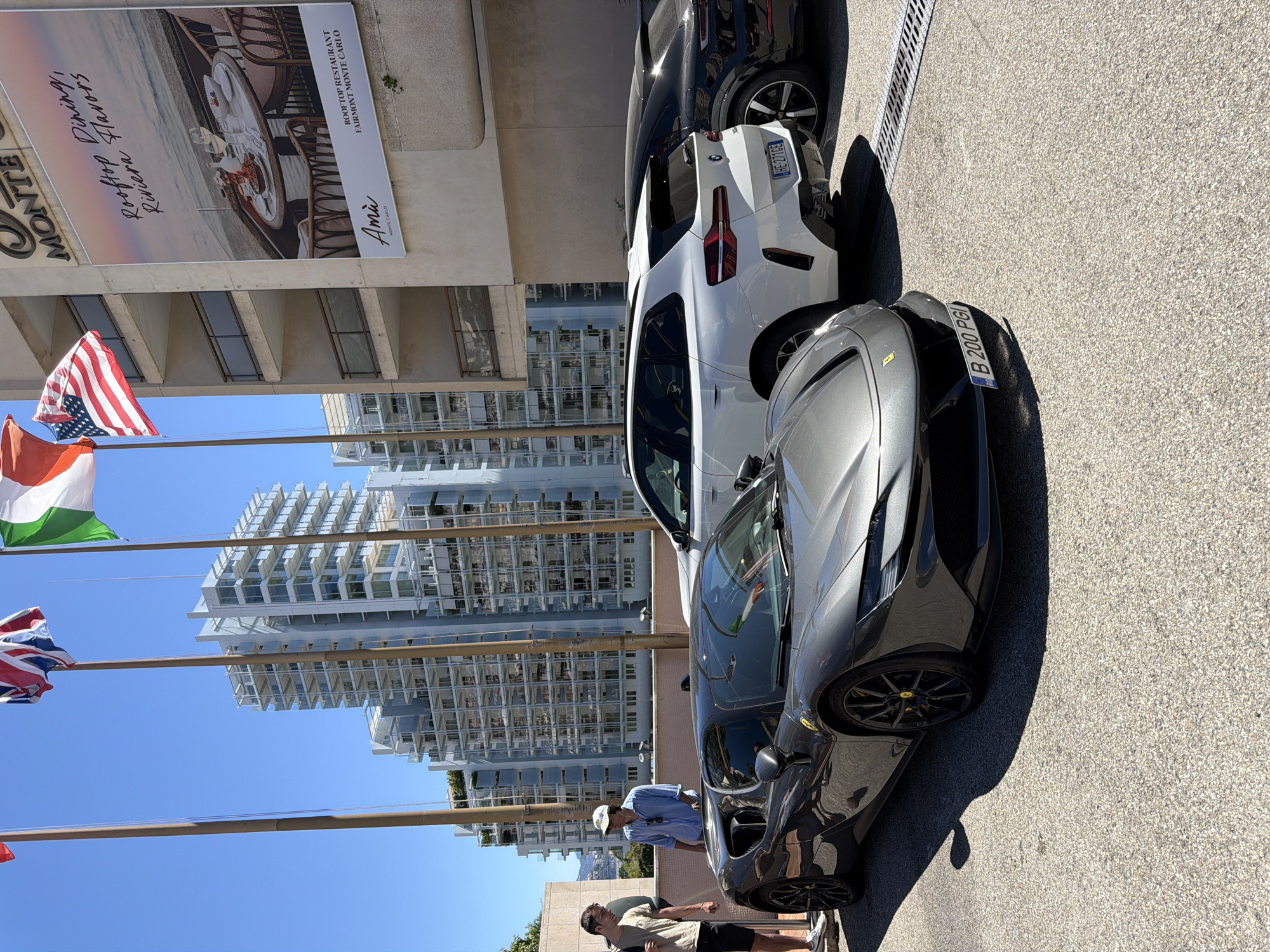 In Monaco, a grey Ferrari SF90 Stradale is parked on asphalt, with a white BMW X3 and a black Porsche Taycan visible behind it. Two young men stand near the Ferrari, one in a light tan shirt and black shorts, the other in a blue button-down shirt and white baseball cap. In the background, a modern white building with balconies and blue-tinted windows rises under a clear blue sky, with several flags, including the Italian and American flags, flying from poles in front of it. A large advertisement for "Rooftop Dining, Riviera Flavors" at Fairmont Monte Carlo is affixed to an adjacent building.