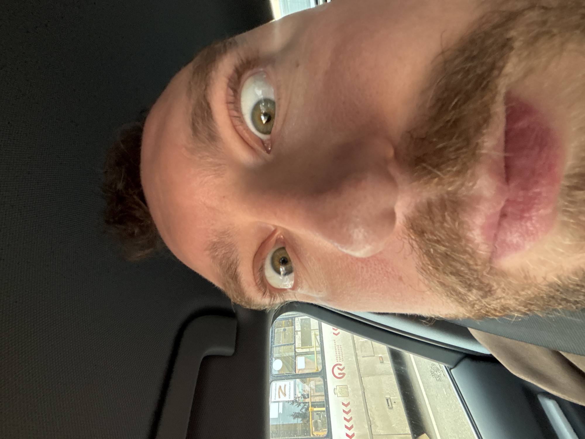 A close-up selfie taken from inside a vehicle. The subject is a man with light skin, hazel eyes, a mustache, and stubble. He appears to be looking upward with an expression of surprise. The view through the car window reveals a Russian bus with a Cyrillic inscription. One can identify a logo for "Krasnoyarsky Gorodskoy Transport" which translates to Krasnoyarsk City Transport. The bus also has a stylized letter "Z" which has become a symbol of the 2022 Russian invasion of Ukraine. Based on the Cyrillic inscription for Krasnoyarsk City Transport, the location of the picture can be identified as Krasnoyarsk, Russia.