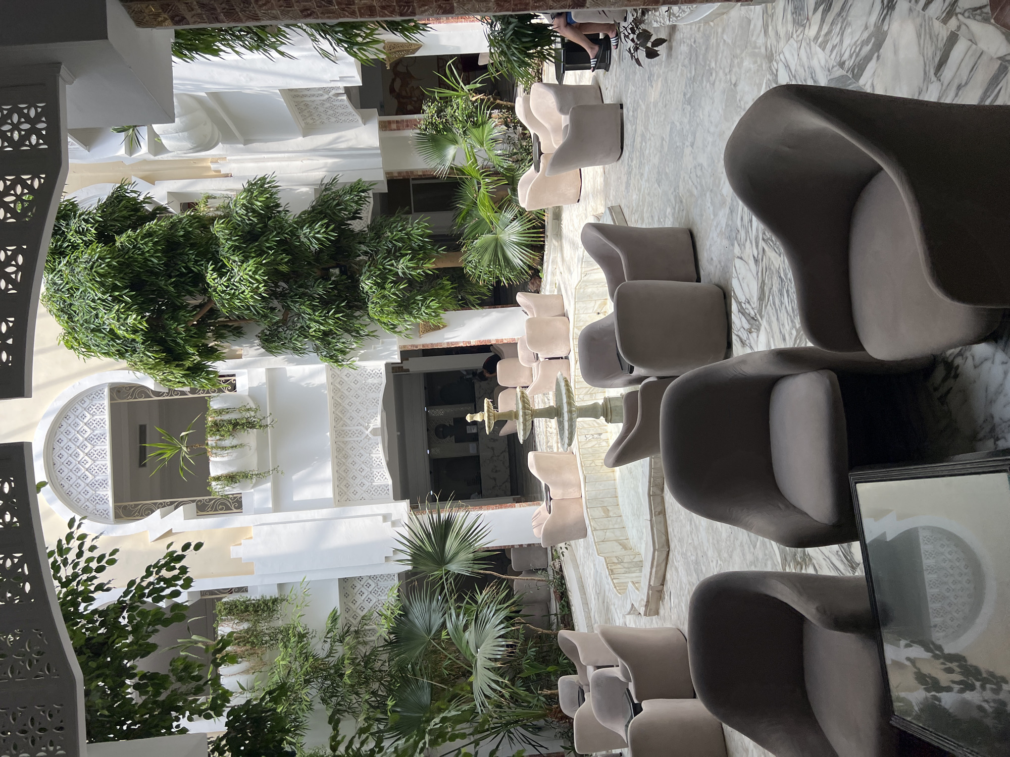 This is a high-angle, eye-level, medium outdoor shot of an open-air atrium in a hotel in Hiboun, Tunisia. The atrium features a central, multi-tiered fountain surrounded by numerous plush, upholstered armchairs in light beige and taupe. Lush green plants, including palm trees and hanging vines, are strategically placed throughout the space, creating a tropical ambiance. The architecture is characterized by white arched windows with intricate geometric cutouts and cream-colored walls. Sunlight filters through the open roof, casting shadows on the marble-tiled floor. In the background, an entrance to the hotel lobby is visible, with a reception desk and people walking. The overall scene conveys a serene and inviting atmosphere, typical of a resort setting.