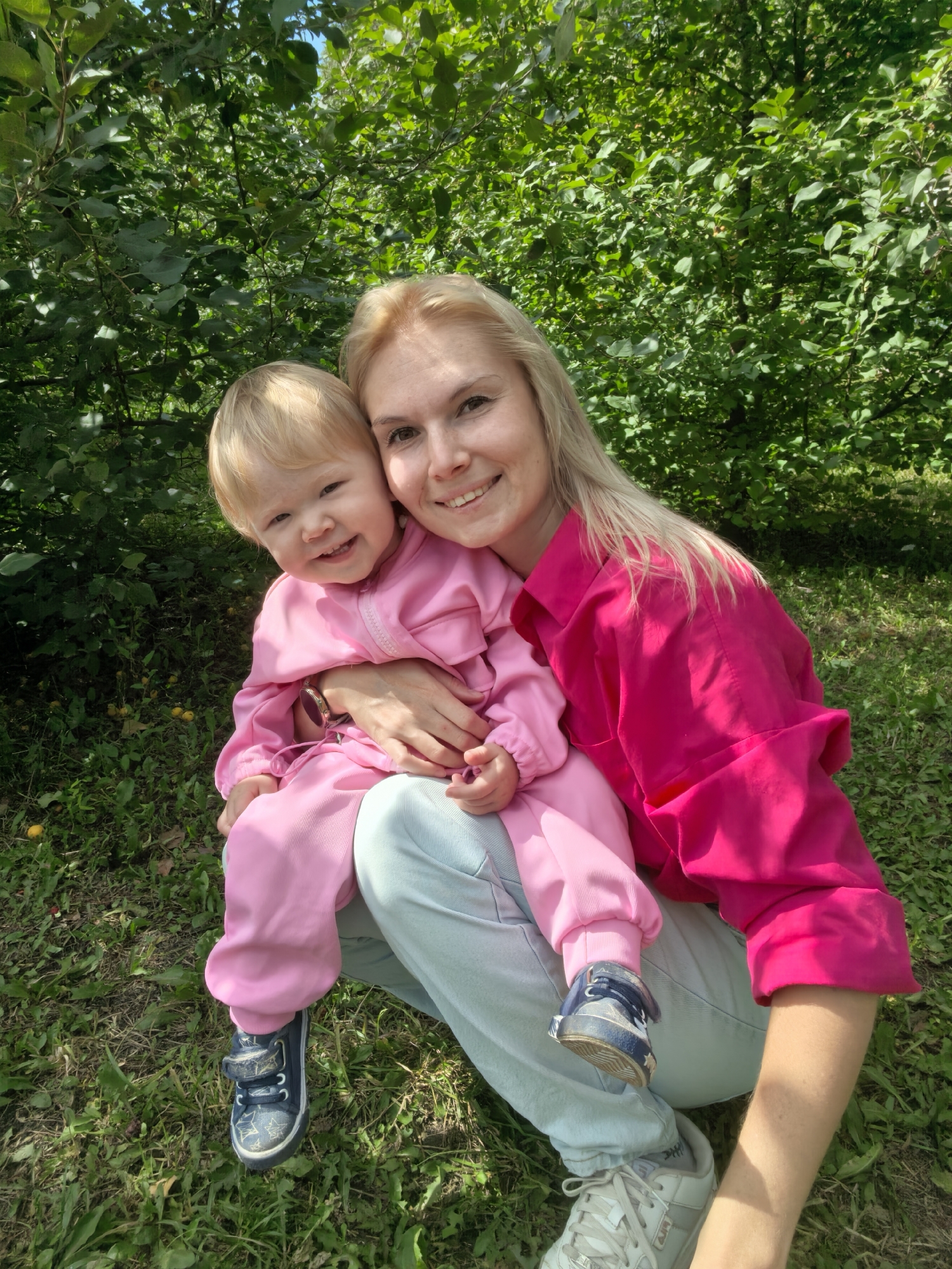 An outdoor, medium-shot photograph captures a blonde woman and a toddler child posing for a picture amidst lush green foliage. The woman, positioned on the right side of the frame, is dressed in a vibrant pink, oversized button-down shirt and light blue pants. She is crouching, holding the toddler, who is on her lap, facing the camera. The child, with blonde hair, is wearing a pink hooded jumpsuit and dark blue sneakers with star patterns. The woman has a warm smile and her arm is around the child. The background is dominated by dense, green leaves from trees and bushes, with patches of grass and some small, yellowish fruits scattered on the ground. The lighting suggests a sunny day. The scene is likely a park or a backyard in Sterlitamak, Russia, given the natural setting.