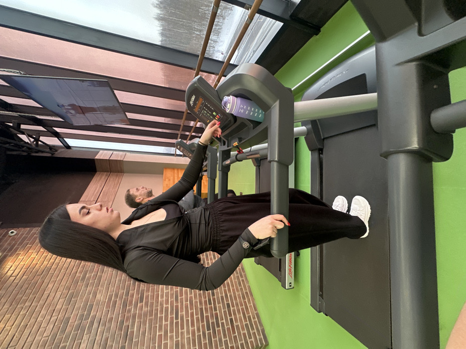 A modern fitness center in Saint Petersburg, Russia, is depicted during daylight hours, characterized by winter conditions visible outside large panoramic windows. A female subject, approximately 20-30 years old, with long dark hair, wearing black athletic attire consisting of a long-sleeved cropped top with a zippered front and button details on the cuffs, a black sports bra, and loose black trousers, along with white athletic shoes, stands on a "Life Fitness" brand treadmill. Her left hand grips the treadmill's front handlebar, while her right hand operates the control panel. A purple-to-teal gradient water bottle rests in a designated holder on the console. A male subject, with short brown hair and a beard, wearing a dark top, is partially visible on an adjacent treadmill further back. The gym interior features a brick-textured wall to the left, a bright green floor, and dark upper walls/ceiling. The exterior view through the large, slanted windows reveals a snow-covered landscape with bare deciduous trees and a road structure, suggesting a peripheral urban or suburban setting within Saint Petersburg. A television screen displaying fitness-related content is mounted on an angled beam near the windows. The scene indicates individuals engaged in or preparing for physical exercise.