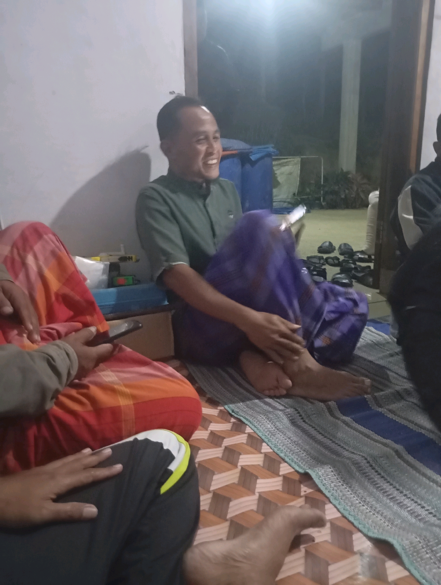 A group of four people are sitting on the floor indoors in what appears to be a dwelling in Ketepung, Indonesia. The room is dimly lit, with a light source casting shadows on the wall. In the foreground, a person wearing a red and orange patterned sarong is holding a smartphone. To their left, another person's hand is resting on dark pants with a white and yellow stripe. In the middle ground, a man wearing a green short-sleeved shirt and a purple sarong is laughing and looking to his left. His legs are crossed, and his bare feet are visible. Behind him, there are several pairs of black sandals neatly arranged on the floor. To the right, another person, partially visible, is wearing a dark jacket with a white shoulder detail. The floor is covered with a woven mat and a tiled area with a geometric pattern. The overall atmosphere suggests a casual gathering.