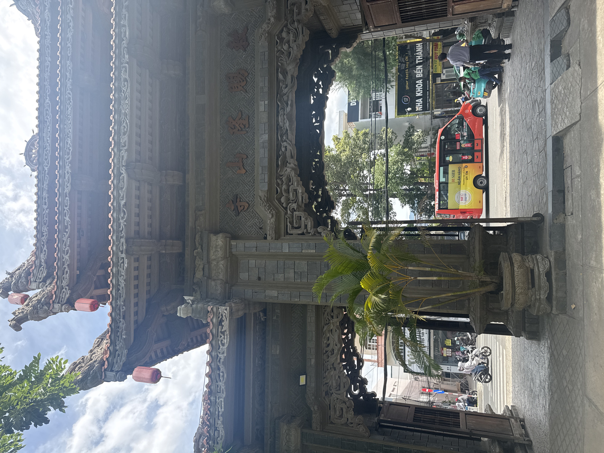 The image captures a street view in an urban setting, with a prominent ancient-style gate in the foreground, featuring intricate wooden carvings and stonework. Red lanterns hang from the gate's roof. Beyond the gate, a street scene unfolds with a red and yellow bus, several motorcycles, and a few pedestrians. Buildings line the street, some displaying signage. A palm tree in a stone pot stands near the gate. The sky is partly cloudy.