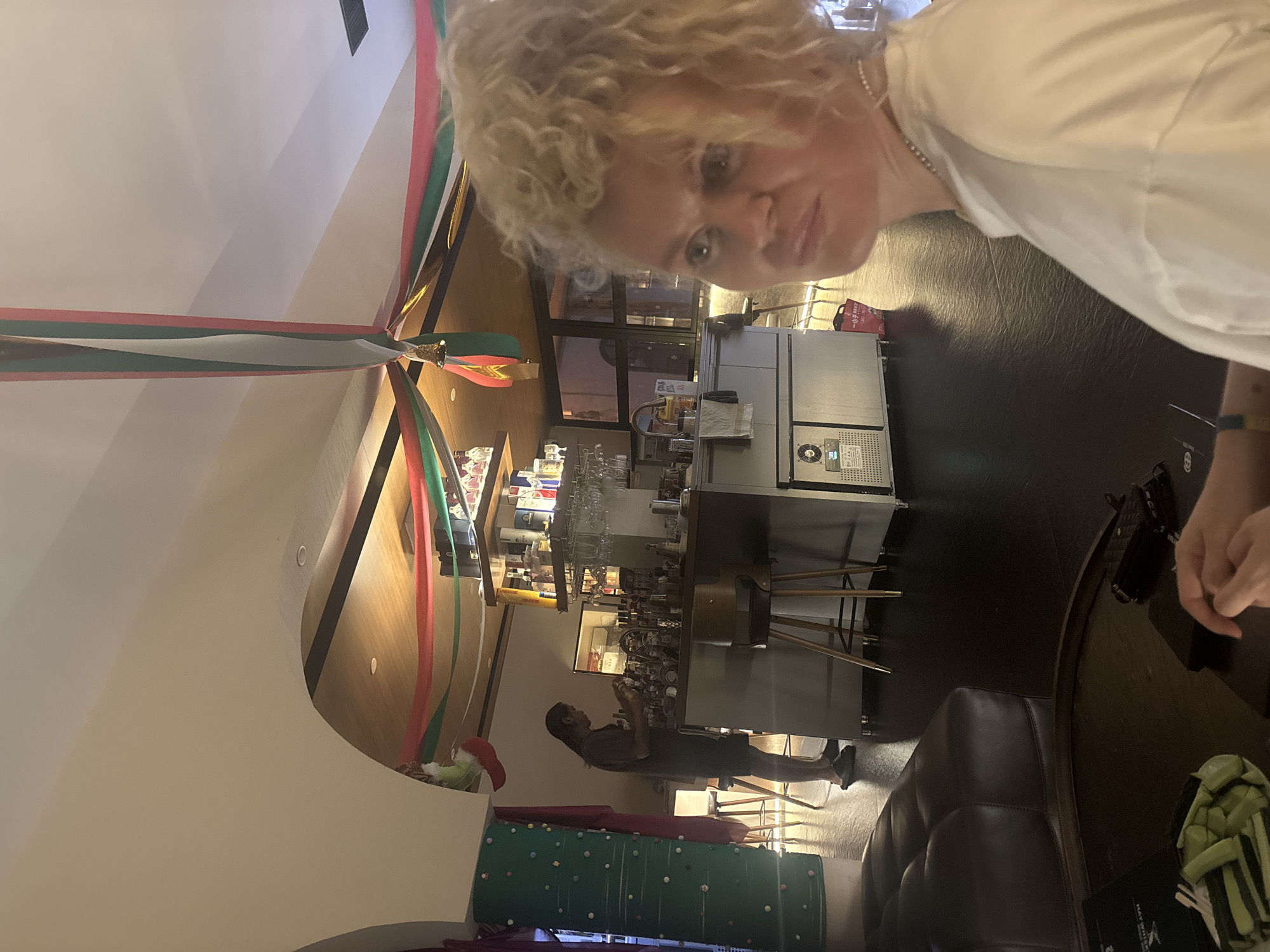 In the dimly lit interior of what appears to be a bar or lounge in Nanhai District, China, a woman with blonde, curly hair and a white shirt is positioned in the foreground, looking slightly to her right with a neutral expression. Behind her, a bar area is visible with a bartender, dressed in dark clothing, attending to bottles and shelves stocked with various beverages. The bar is illuminated by warm, overhead lighting, and colorful ribbons in red, green, and white are draped across the ceiling, suggesting a festive decoration. A dark leather sofa and a round wooden table with a plate of sliced green vegetables and a black object are in the lower-left portion of the frame, indicating a seating area. The floor is dark and reflective.