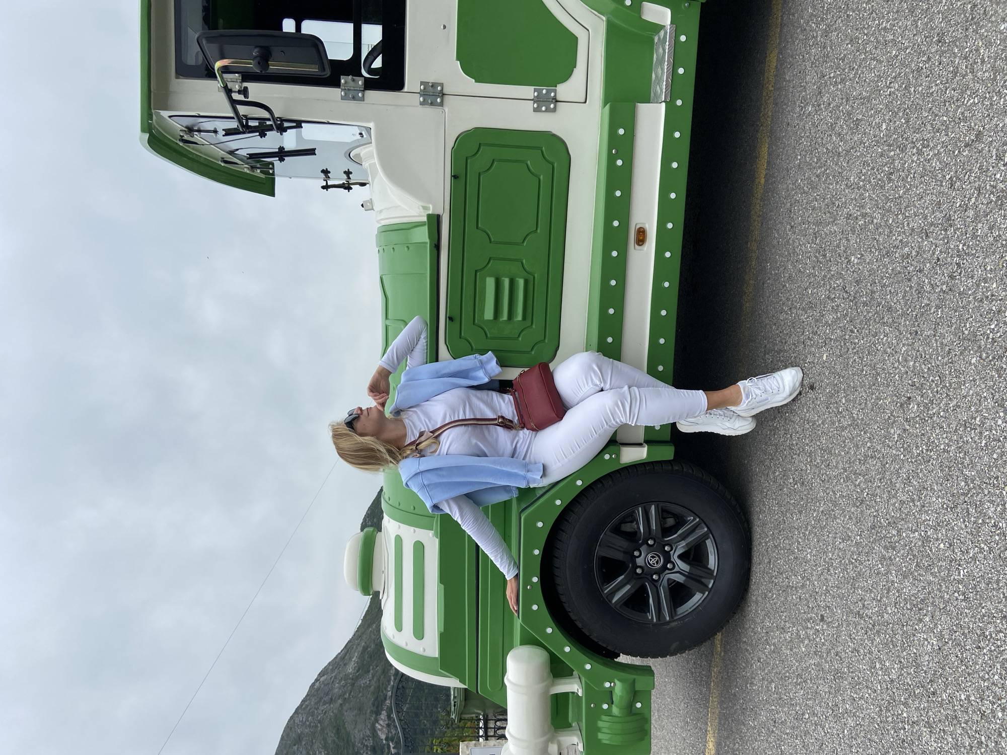 A full-length photograph depicts a blonde woman lying on the side of a green and white tourist vehicle, likely in Lipa Dobrska, Montenegro, considering the mountainous background. The woman is wearing a white long-sleeve shirt, white pants, white sneakers, a light blue jacket, and sunglasses. A small red purse is slung across her torso. She is positioned on the vehicle's side, with part of her body resting on the front tire. The vehicle is predominantly green with white accents and circular details. The background consists of a gray asphalt surface and a sky with some clouds, in addition to a mountain in the distance.