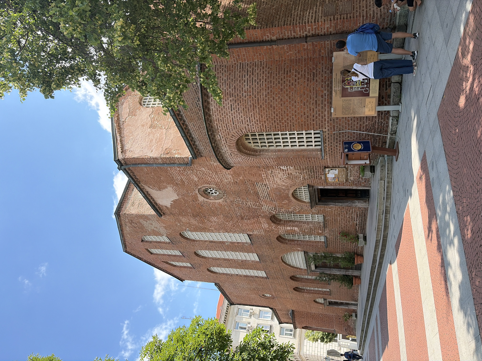 The image depicts the Church of St. Sophia in Sofia, Bulgaria, a historic red brick building with arched windows and a rounded apse. The church is situated in a public square paved with red and gray brick patterns, with some areas showing dappled sunlight and shadows.
In the foreground, two people, a woman wearing a white top and striped trousers and a man in a blue t-shirt and shorts, are standing and looking at an informational display board. The man is holding a phone up, possibly taking a picture. To their right, another person, wearing dark shorts and a t-shirt, is seated on the ground, also holding a phone.
To the left of the church entrance, two potted evergreen trees and a smaller potted plant are visible. A person in a dark blue shirt and dark trousers is walking away from the church entrance in the distance. Behind the church, a modern building with white facades and windows can be seen. Lush green trees with some light-colored blossoms partially obscure the sky and the upper sections of the church. The sky is blue with scattered white clouds.