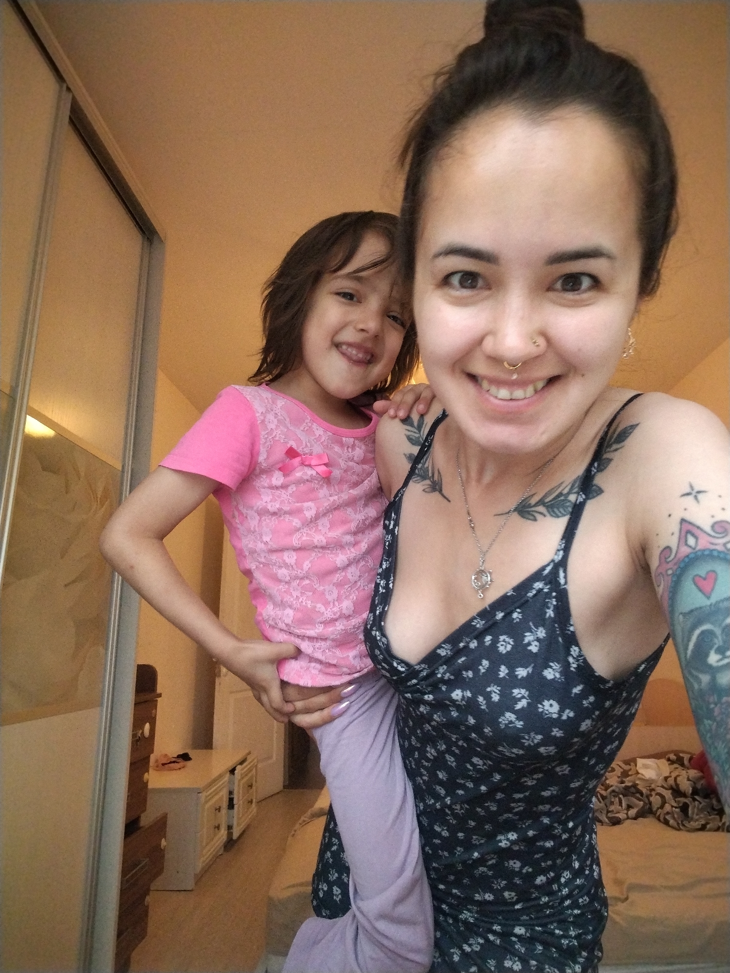A woman is taking a selfie while holding a young girl. The woman has dark hair pulled up in a bun, and is wearing a dark floral print dress and a silver necklace. She has tattoos on her chest and arm, as well as a nose ring. The girl is wearing a pink t-shirt with a lace overlay and a pink bow. She has brown hair and is smiling. They appear to be indoors, in what looks like a bedroom. Behind them, a large mirrored closet door is visible, and further in the background, a dresser and a bed can be seen. The lighting suggests it might be daytime. The overall mood is happy and affectionate.