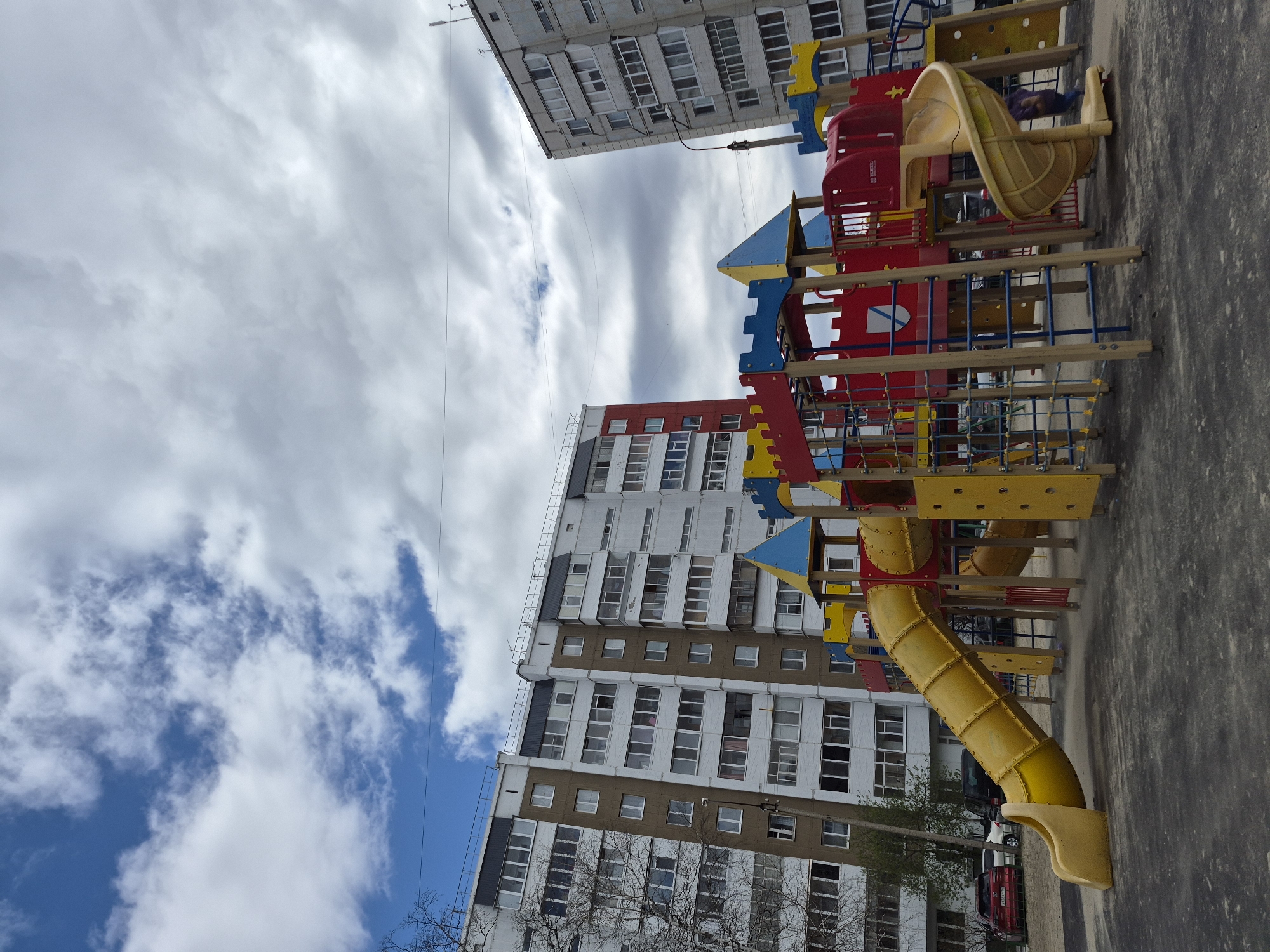 Here is a description of the image:
The image shows a brightly colored playground in an urban setting. The playground features a red and yellow structure resembling a castle, complete with slides and climbing areas. The background includes tall, multi-story apartment buildings, common in Russian cities. The sky is filled with a mix of clouds and blue patches. A few trees are visible. The image appears to be taken outdoors. Although not directly confirmed in the image, the presence of Russian architecture and infrastructure hints towards the location as Tobolsk, Russia.