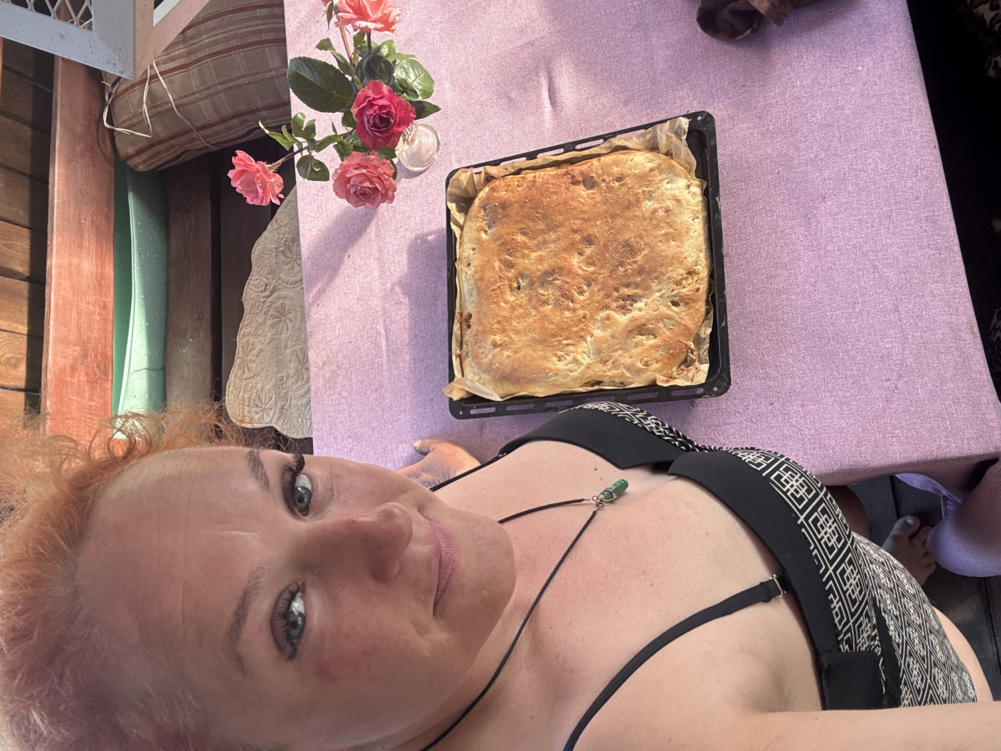 The image shows a woman with red hair and blue eyes, wearing a black and white patterned bikini top and a necklace with a green pendant. She is looking at the camera and smiling. In front of her is a table covered with a pink tablecloth, on which rests a baked dish with bread or a similar pastry and a small vase with pink roses. The background shows wooden structures and what appears to be outdoor seating. The scene is set outdoors. The provided metadata indicates that the image was taken in Chaikovsky, Russia.