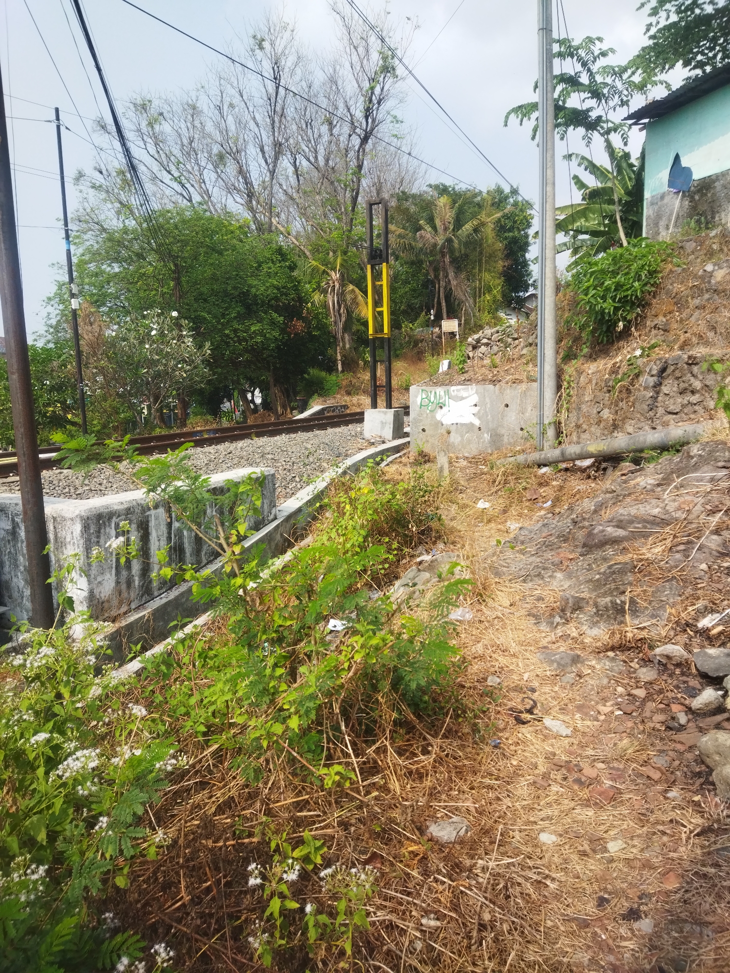 The image depicts a rural railway track area in Giripurwo, Indonesia. A double railway track runs horizontally across the middle ground, flanked by ballast. To the left of the tracks, a concrete retaining wall with some weathering and graffiti is visible, bordered by green vegetation and some white flowering plants in the foreground. Behind the wall and tracks, a dense line of trees and vegetation forms a natural backdrop, with a bright yellow and black railway signal pole situated prominently. To the right of the tracks, a slope covered in dry grass, rocks, and some debris leads up to a stone wall supporting a building with a light blue wall and dark roof. A cylindrical pipe runs along the slope. The sky is overcast. No people are present in the scene.