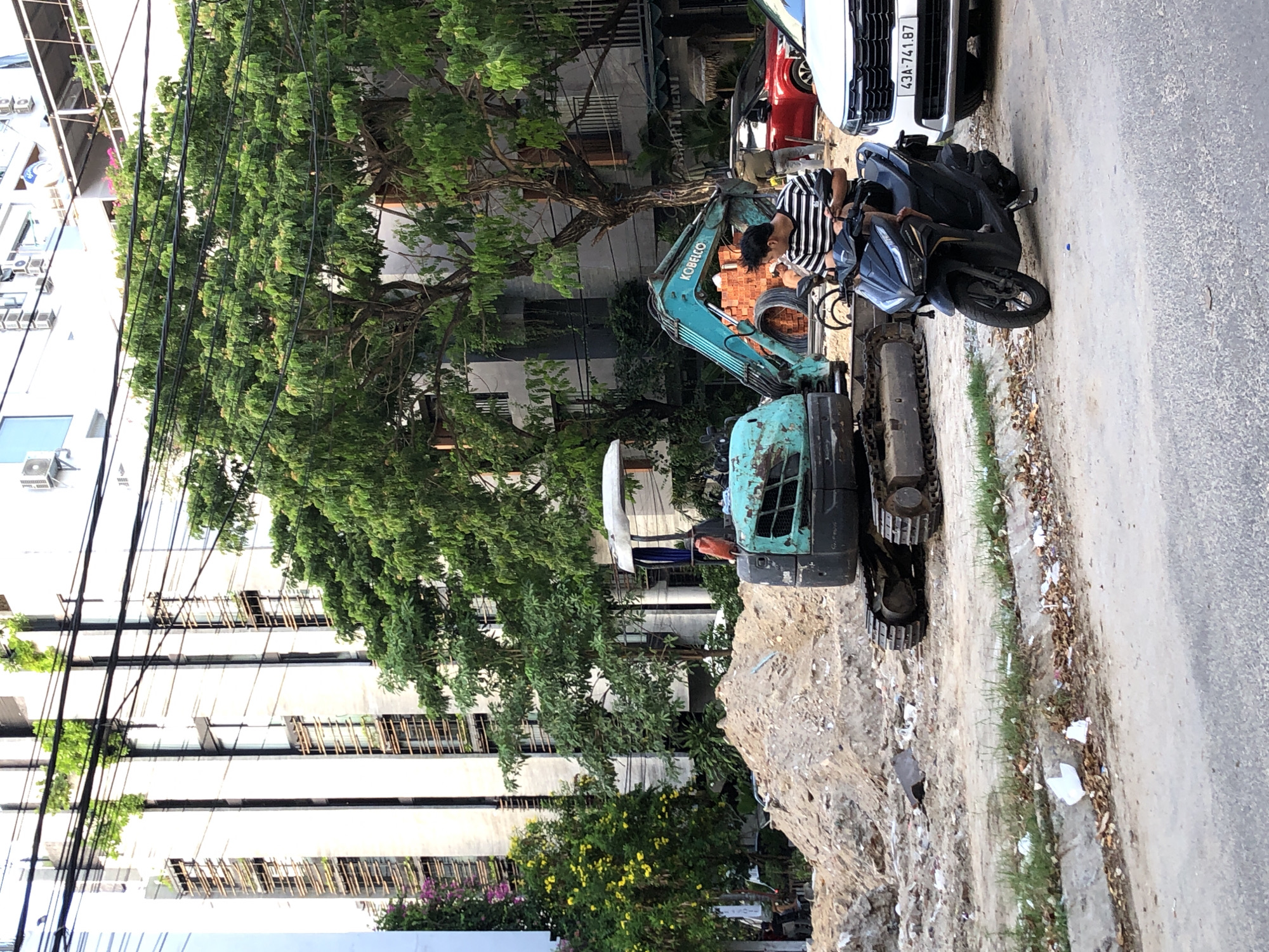 An outdoor scene in Đà Nẵng, Vietnam, depicts a construction site with two excavators and a pile of dirt. In the foreground, a person is riding a black motorcycle. To the right of the motorcycle, a white car is parked. In the background, there is a multi-story building with balconies and vegetation. Power lines are visible crisscrossing the upper portion of the image.