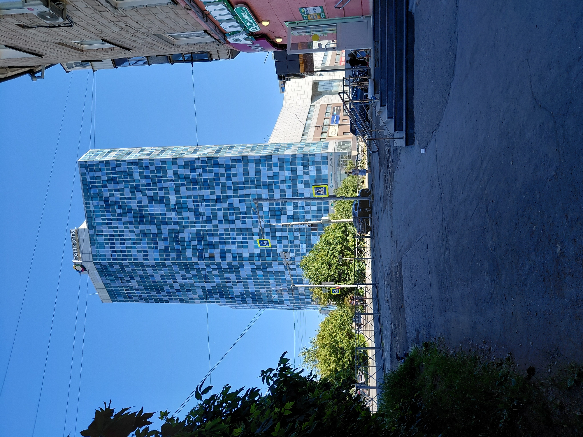 The image captures a street scene in a city, likely on a clear, sunny day. Dominating the skyline is a modern, multi-story building with a facade composed of blue and white checkered glass panels. The building's apex features a sign with the Cyrillic text "САН СИТИ" and a colorful logo. To the right of this skyscraper, a brick building with windows and an air conditioning unit is visible. In the foreground, a paved area with some cracks leads towards the entrance of a pink building, where stairs with handrails are present. Several people are seen near the entrance of the pink building and on the street in the mid-ground. Cars are parked and in motion on the street behind a metal fence. Lush green trees and foliage are interspersed throughout the scene, with a cluster of leaves in the upper left corner partially obscuring the view. Yellow pedestrian crossing signs are placed on poles near the street. The overall environment suggests an urban setting with a mix of commercial and residential elements.