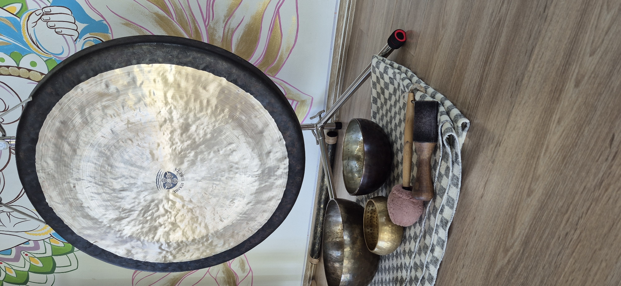 The image shows a gong hanging on a stand, with a variety of mallets and singing bowls beside it. The gong is circular, with a dark rim and a reflective central surface. It has a brand or logo on the lower part of the reflective surface. The stand appears to be metal and adjustable. Next to the gong, hanging on the stand, is a dishtowel with a gray and white checkerboard pattern. There are several singing bowls of varying sizes and colors, alongside different mallets used to strike the bowls or gong. The background includes a wall with a colorful abstract pattern. The location is Unknown, Unknown.