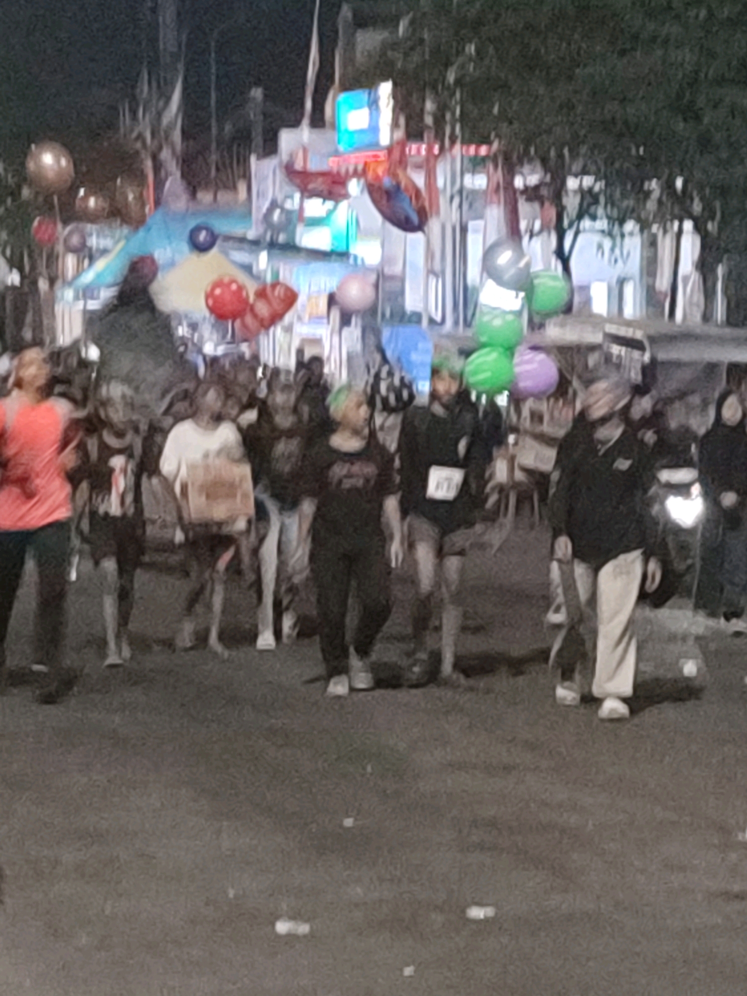 The image depicts a nighttime street scene in Jember, Indonesia, likely during a festival or event. A group of people are walking on a paved surface, appearing to be in a procession or parade. The individuals are varied in age and attire, with some wearing t-shirts, shorts, and long pants. There are balloons visible in the background, some of which are large and spherical, in various colors including red, gold, and green. In the middle ground, there are structures that appear to be temporary stalls or decorations, illuminated by lights. The overall atmosphere is festive, with a sense of community gathering. The presence of a motorcycle with its headlight on suggests active movement or a waiting vehicle within the scene. The lighting is dim, characteristic of an evening or nighttime setting.