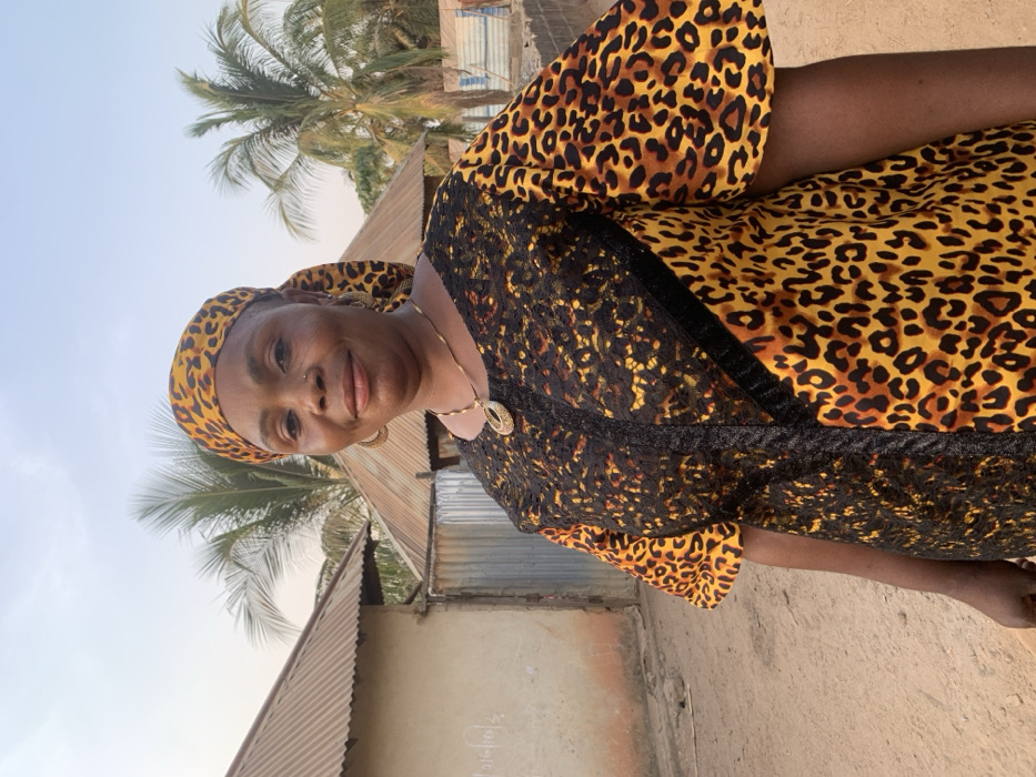 An adult Black woman wearing a leopard print headscarf and dress stands outside. Her dress is leopard print with a black lace overlay on the front. She is also wearing a gold necklace and earrings. A corrugated metal roof, a concrete wall, and a palm tree are visible behind her. The sky is light blue and the ground is tan. The photo is slightly tilted to the left.