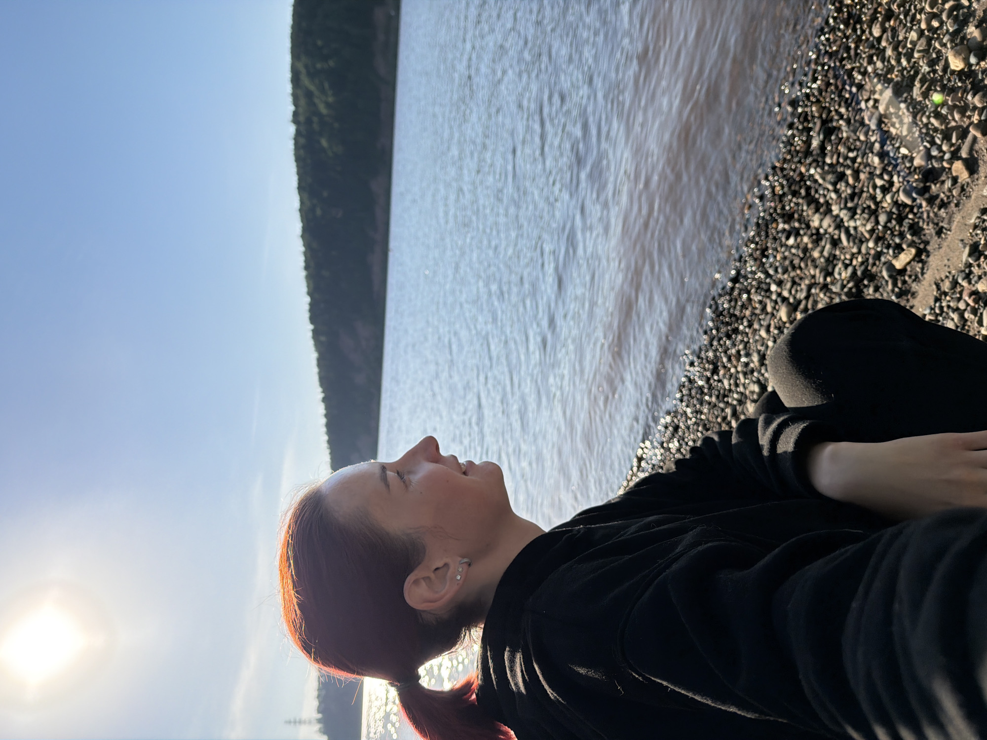A medium-angle shot of a woman sitting on a rocky beach near Noviy, Russia. The woman has red hair and is wearing a black sweatshirt and pants. She is looking up at the sky, which is a clear blue. The sun is shining brightly in the bottom left corner. In the background is a body of water, and beyond that, a hillside covered in green trees. The beach consists of small, dark-colored pebbles.