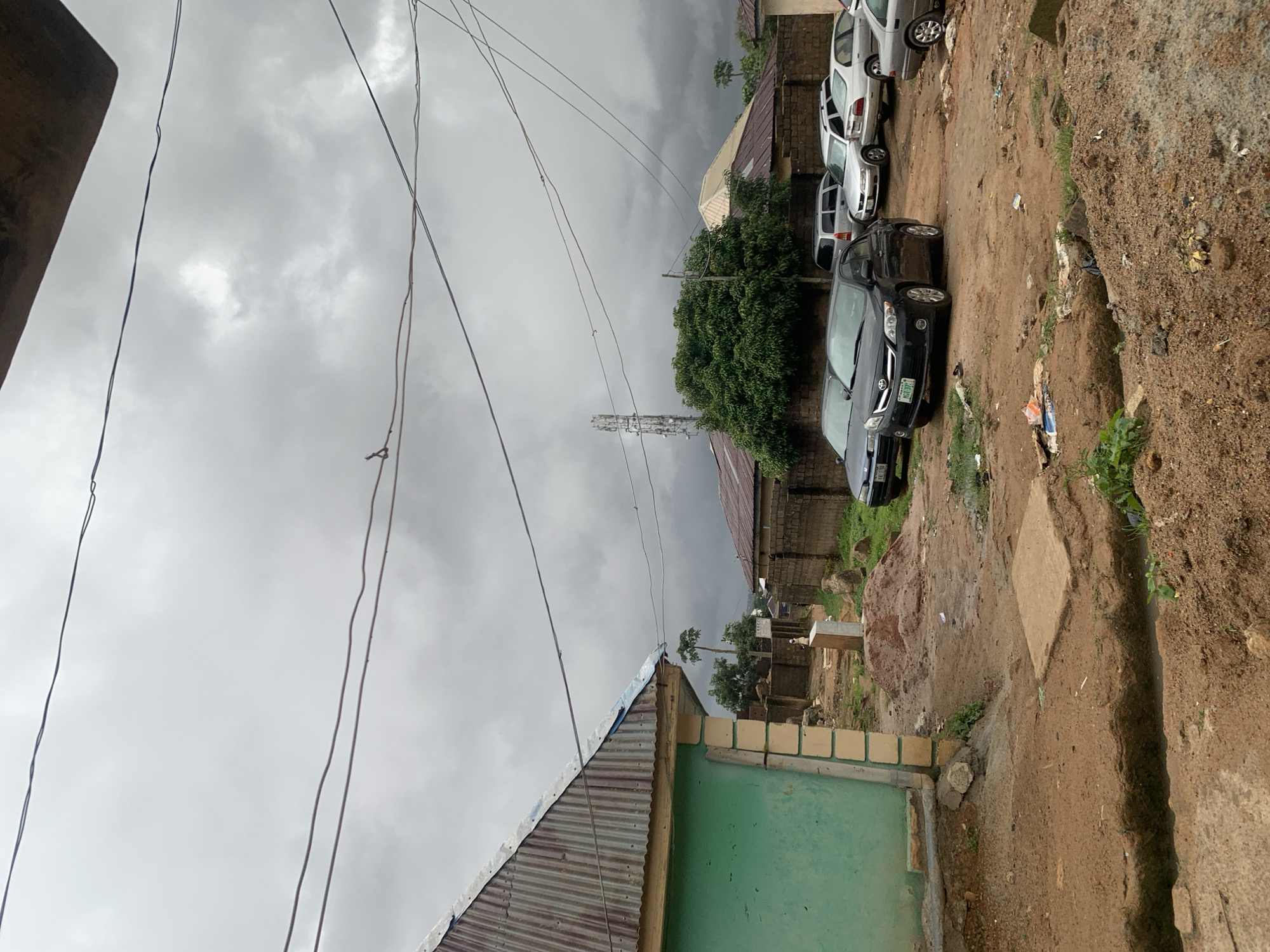 The image depicts an overcast day in a residential area of Jos, Nigeria. In the foreground, a dirt road with a trench runs across it. To the left, a partially visible green building with a corrugated metal roof stands prominently. Several cars are parked on the dirt lot, including a dark sedan with a license plate reading "XCT 310". In the background, more buildings with corrugated roofs and brick walls are visible, along with a telecommunications tower. The environment appears somewhat unkempt, with scattered debris and piles of dirt. The sky is a dull gray, suggesting impending rain.