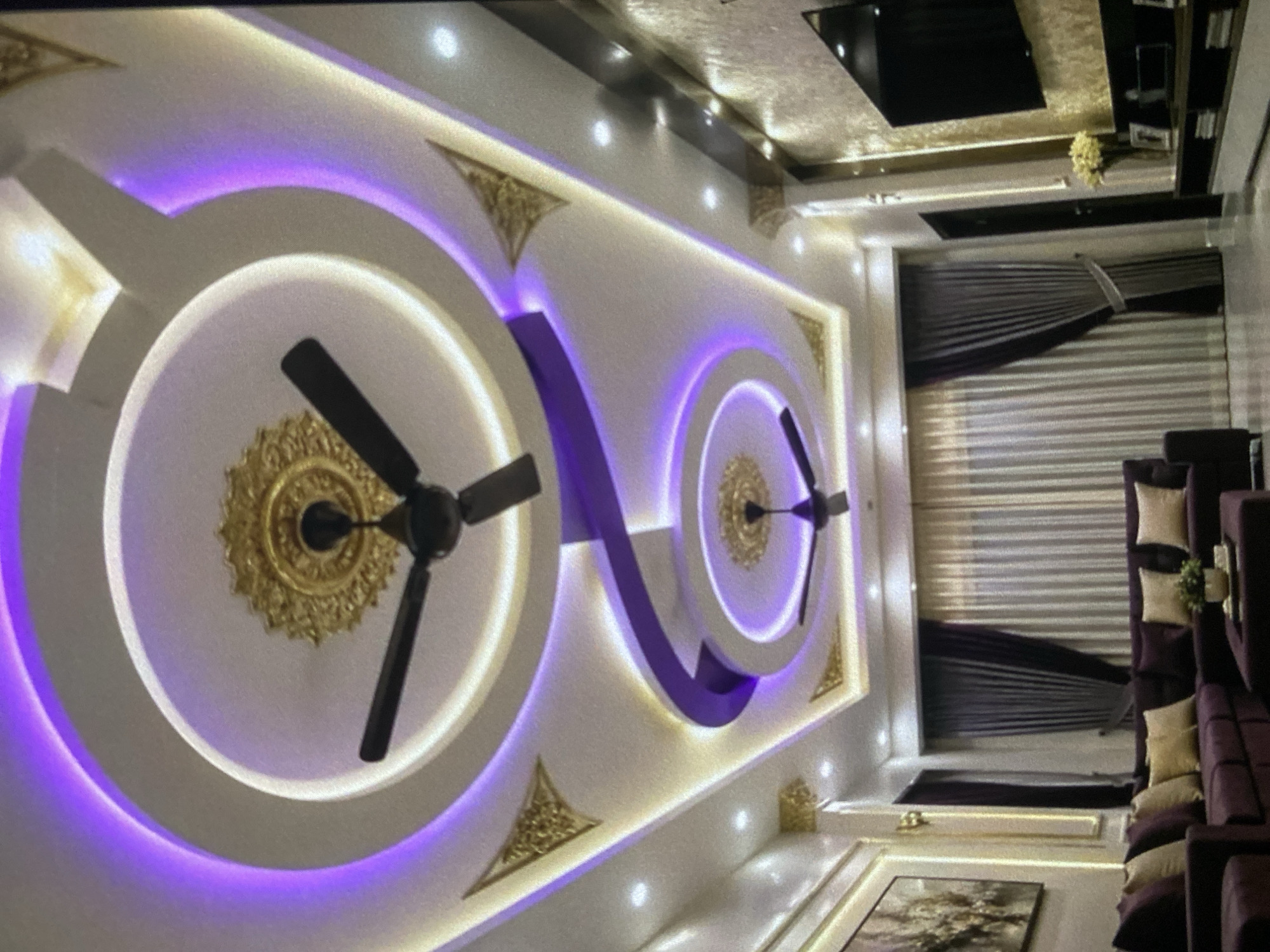 A brightly lit interior view, likely in Lhokseumawe, Indonesia, showcases an ornate ceiling design. The primary focus is on two circular recesses with decorative gold centers, each featuring a black ceiling fan. These recesses are bordered by concentric white rings illuminated with purple lighting. Gilt triangular embellishments appear at various points along the ceiling's edge. The lighting suggests a modern, upscale ambiance. Further into the image is a room setting including brown striped curtains, a dark colored sectional sofa with cream colored accent pillows, and a framed picture on the wall. A black rectangular television is mounted above near the ceiling.