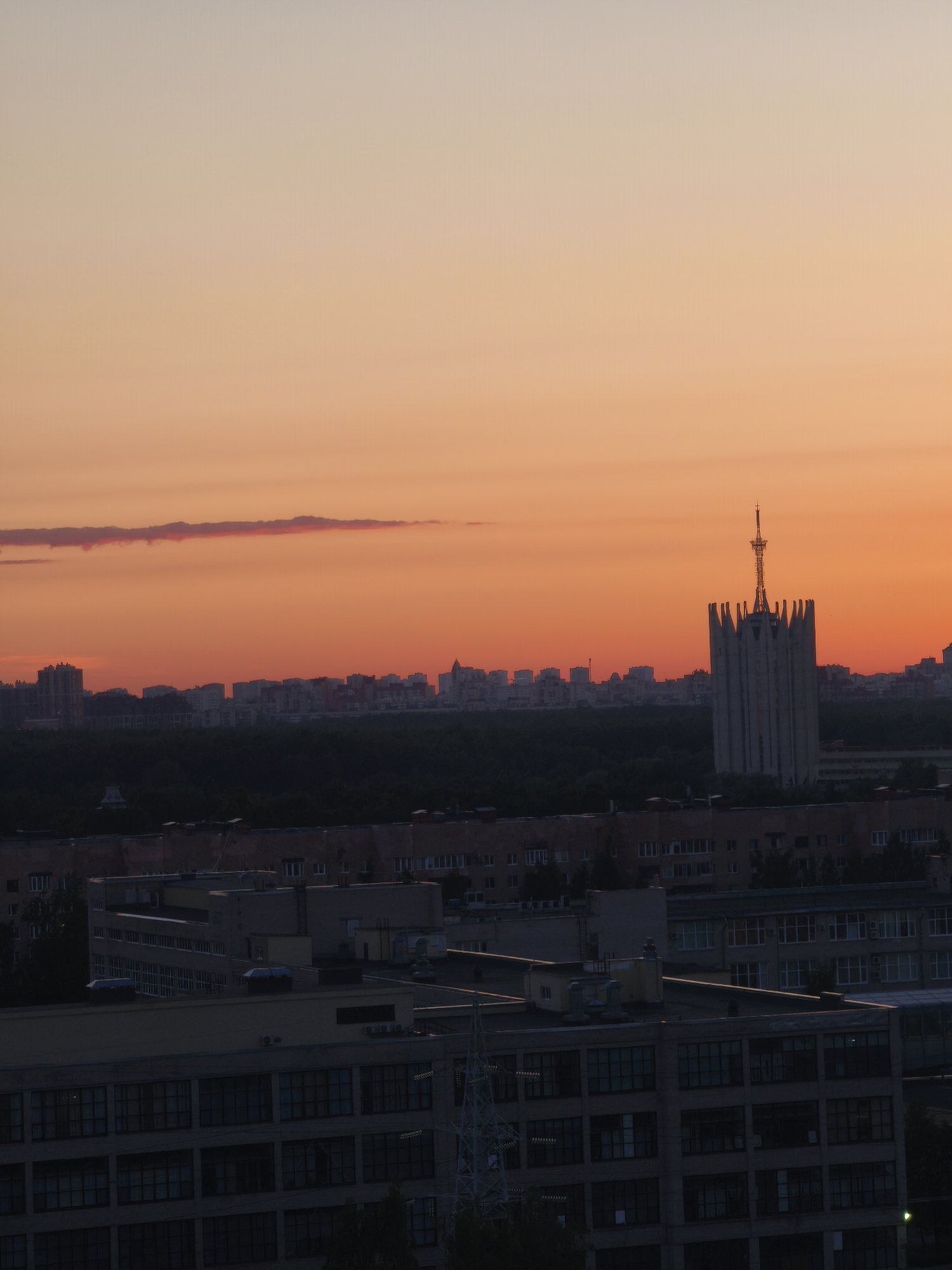 The image displays an urban landscape at sunset, characterized by a sky featuring a gradient of orange, red, and pale yellow hues, with a single elongated, thin cloud stretching horizontally in the upper left quadrant. The distant horizon reveals a city skyline composed of numerous multi-story buildings. Prominently situated on the right side of this distant skyline is a tall, light-colored building with a distinctive architectural crown of pointed, vertical elements and a prominent antenna spire. This building is identifiable as the Presidium of the Russian Academy of Sciences. Below the distant cityscape, a dense, dark band of trees or forest occupies the mid-ground. In the foreground, several multi-story buildings, appearing largely silhouetted and dark against the bright sky, feature numerous windows in a grid pattern. A partial view of a grey metal lattice structure is visible on the far lower left. No individuals or specific actions are depicted within the scene. The location is Moscow, Russia.