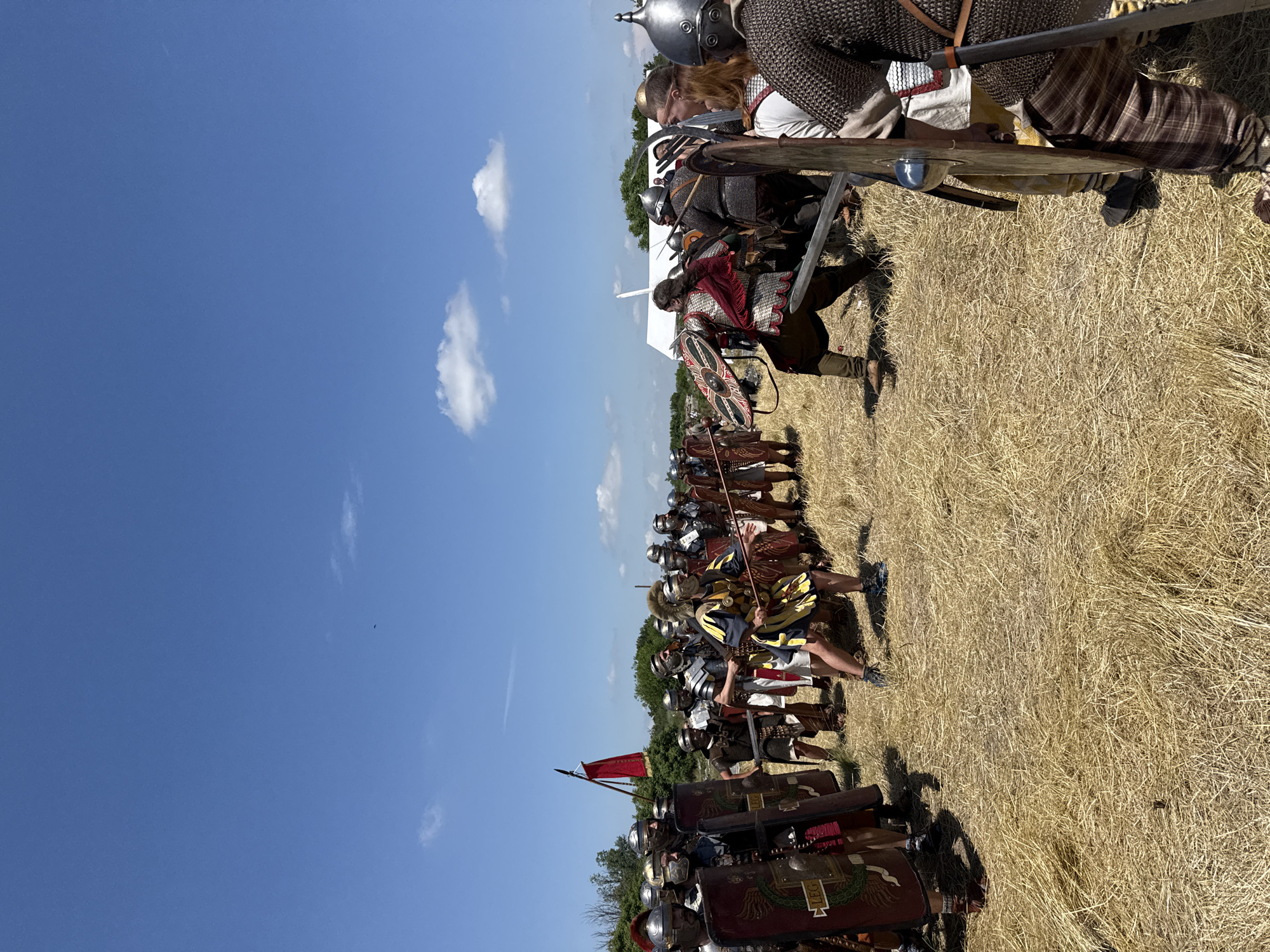 The image captures an outdoor historical reenactment of a battle scene in Gigen, Bulgaria, under a clear blue sky with sparse cumulus clouds. The foreground and mid-ground consist of a field covered in dry, golden straw. Two distinct groups of participants, dressed in historical military attire, are positioned facing each other.
The group on the left, primarily in the left half of the frame, is costumed as Roman soldiers. They wear helmets, segmented armor (lorica segmentata or hamata), and carry large rectangular red shields (scuta) emblazoned with gold or green markings including "LEG." Several members hold spears (pila). One central figure within this group wears a distinctive yellow and blue tunic with a plumed helmet, also holding a spear.
The group on the right, extending towards the right edge of the frame, is attired in chainmail, helmets, and carries round shields. Members are depicted holding swords.
In the center of the image, two figures are engaged in simulated close-quarters combat. One figure, associated with the Roman-style group, wears scale armor (lorica squamata) with a red cloak, a helmet, and holds a sword with a round shield. This figure is in a crouched combat stance, facing right. The opposing figure, from the barbarian-style group, wears chainmail and a helmet, wielding a sword with a visible round shield.
In the distant background to the left, green foliage and small trees are visible, with a red banner atop a pole. To the right in the distant background, a white tent structure is present. The scene is illuminated by bright daylight.