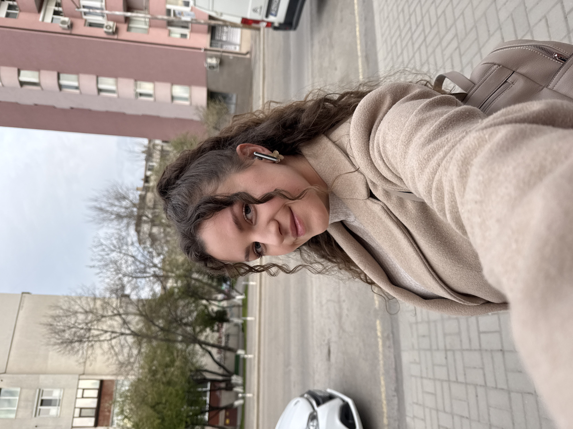 A young woman with curly brown hair is posing for a selfie on a city street. She is wearing a beige coat and has a beige backpack over her shoulder. An earbud is visible in her left ear.
Behind her, a street stretches out with buildings on either side. On the left, there is a pink apartment building. On the right, the sidewalk is paved with square grey tiles. Trees with bare branches are also visible. A white car is visible at the bottom of the frame.
The location is probably Sofia, Bulgaria.