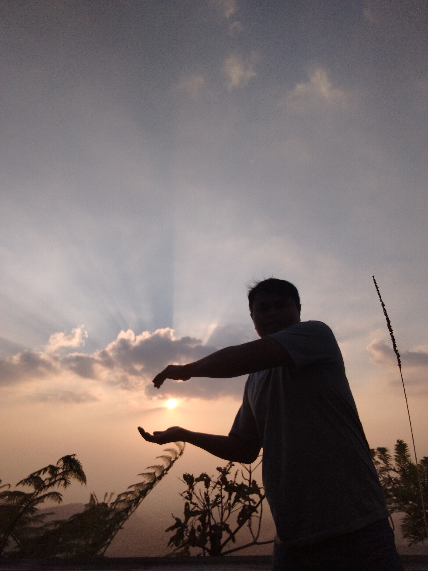 In Gumeng, Indonesia, a person stands silhouetted against a sky illuminated by crepuscular rays emanating from the setting sun. Their right arm is extended, hand cupped, as if holding the sun. Their left arm is raised, hand open, positioned above the right hand. The person is wearing a t-shirt and appears to be standing on an elevated surface, possibly a balcony or rooftop, with vegetation visible in the foreground. The sky is a gradient of soft oranges and purples, with scattered clouds.