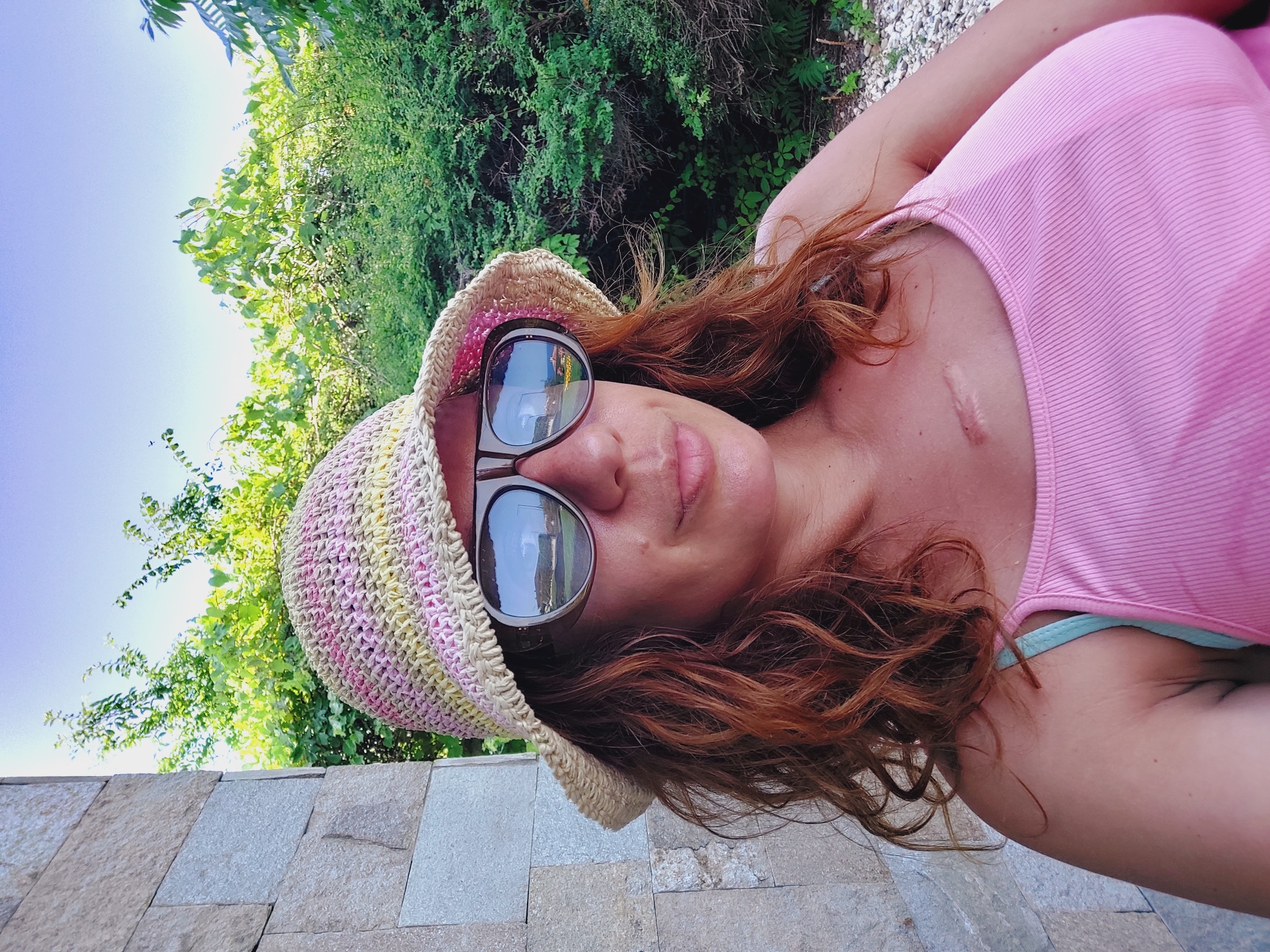 In Sozopol, Bulgaria, a woman with reddish-brown wavy hair, wearing a pink and yellow striped straw hat and oversized sunglasses, is captured in a selfie. She is wearing a pink ribbed tank top and a light blue bra strap is visible. A scar is present on her chest, near her collarbone. The background features a stone wall to the left and lush green foliage and trees under a clear blue sky. The sunglasses reflect a landscape with a road, buildings, and vegetation, possibly indicating the woman is outdoors in a scenic area.